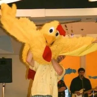 Person in yellow chicken costume being lifted in the air with a band playing in the background.
