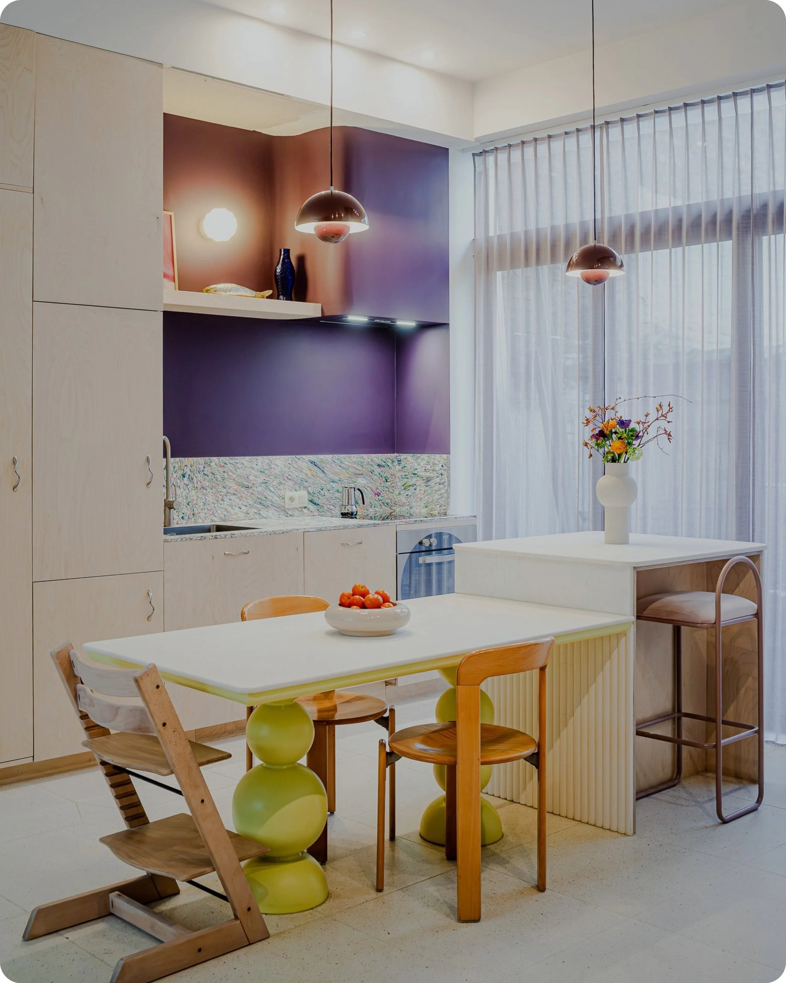 Cuisine de Maison Savoie avec un comptoir en plastique recyclé, des armoires en bois clair, un mur violet, une table à manger blanche avec des chaises en bois, une lampe suspendue, une grande fenêtre avec des voiles blancs et un vase avec des fleurs.