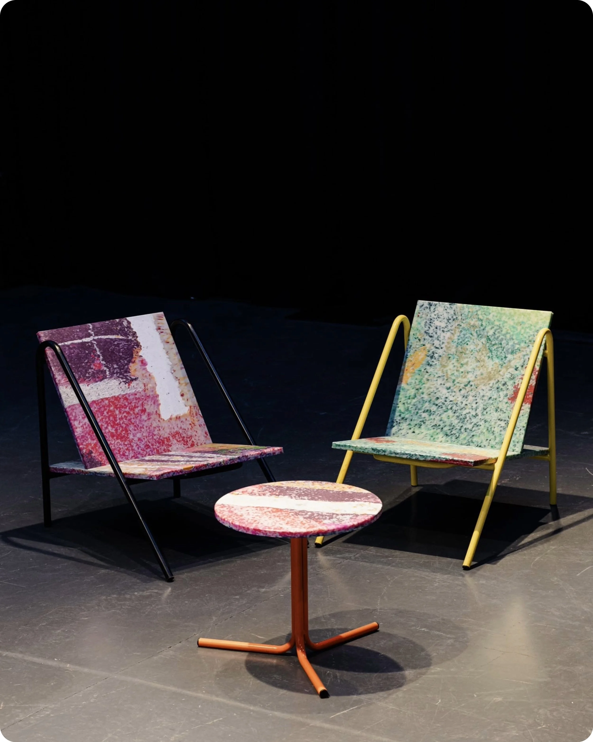 Vue de face de deux fauteuils d'Ateliers J&J et petite table ronde au milieu, le tout éclairé d'au dessus avec fond noir, meubles réalisés en plastique recyclé HDPE de plein de couleurs vives.