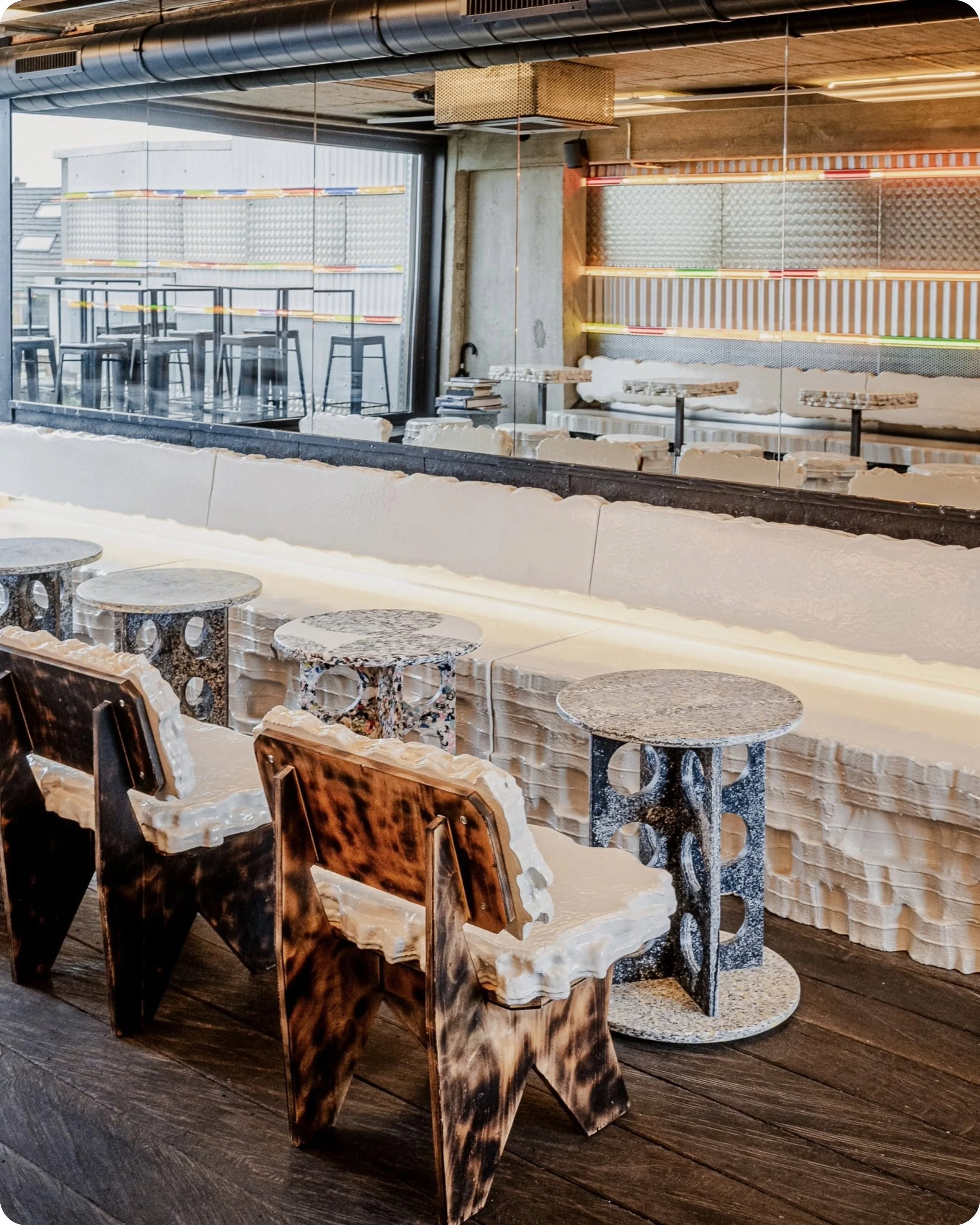 Intérieur d'un bar qui est le "Rooftop Perché" au Jam Hotel, avec des tables en plastique recyclé de divers designs, des chaises en bois et en plastique, ouverture sur une terrasse extérieure avec des tabourets hauts, murs en béton, décor artistique
