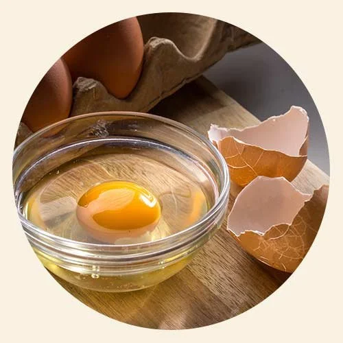 A raw egg cracked into a glass bowl next to broken eggshells on a wooden surface