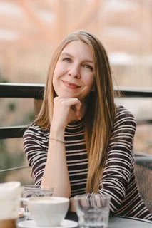 Young woman with long blonde hair wearing a striped shirt, smiling and resting her chin on her hand at a table with drinks