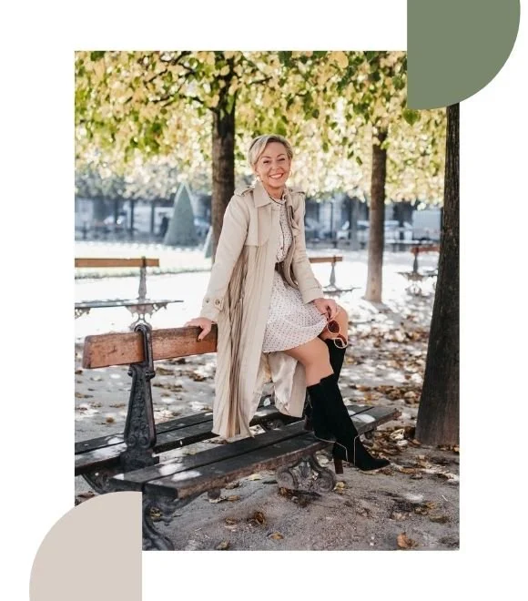 A smiling woman sitting on a park bench among trees with fall foliage, wearing a beige trench coat, white dress, and black knee-high boots.