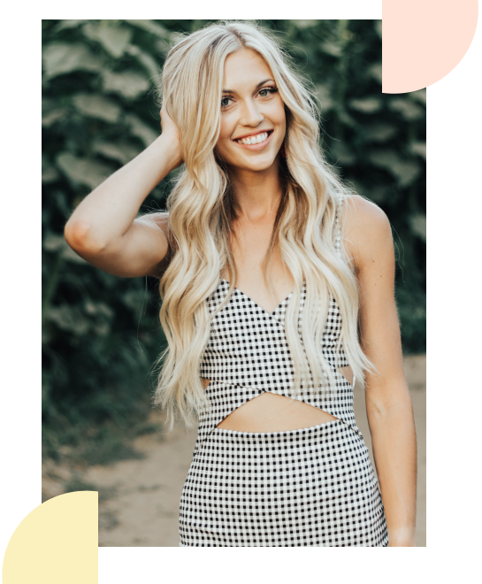 A young woman with long blonde hair wearing a sleeveless, black-and-white checkered crop top and matching skirt, smiling outdoors with green foliage in the background.