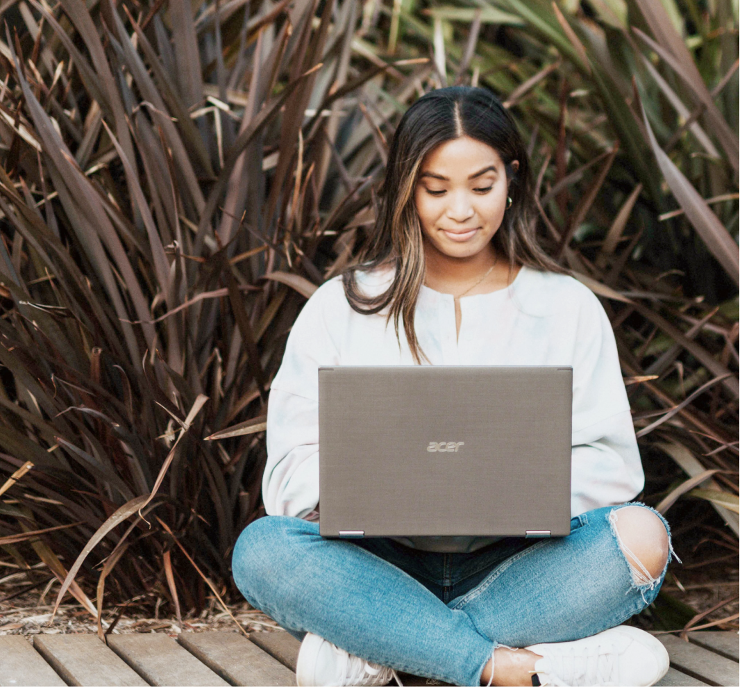 Young woman sitting cross-legged on wooden deck, using a laptop outdoors with tall plants in the background.