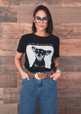 A woman with dark hair and glasses wearing a black T-shirt with a dog graphic and text, tucked into high-waisted blue jeans, standing in front of a wooden wall.