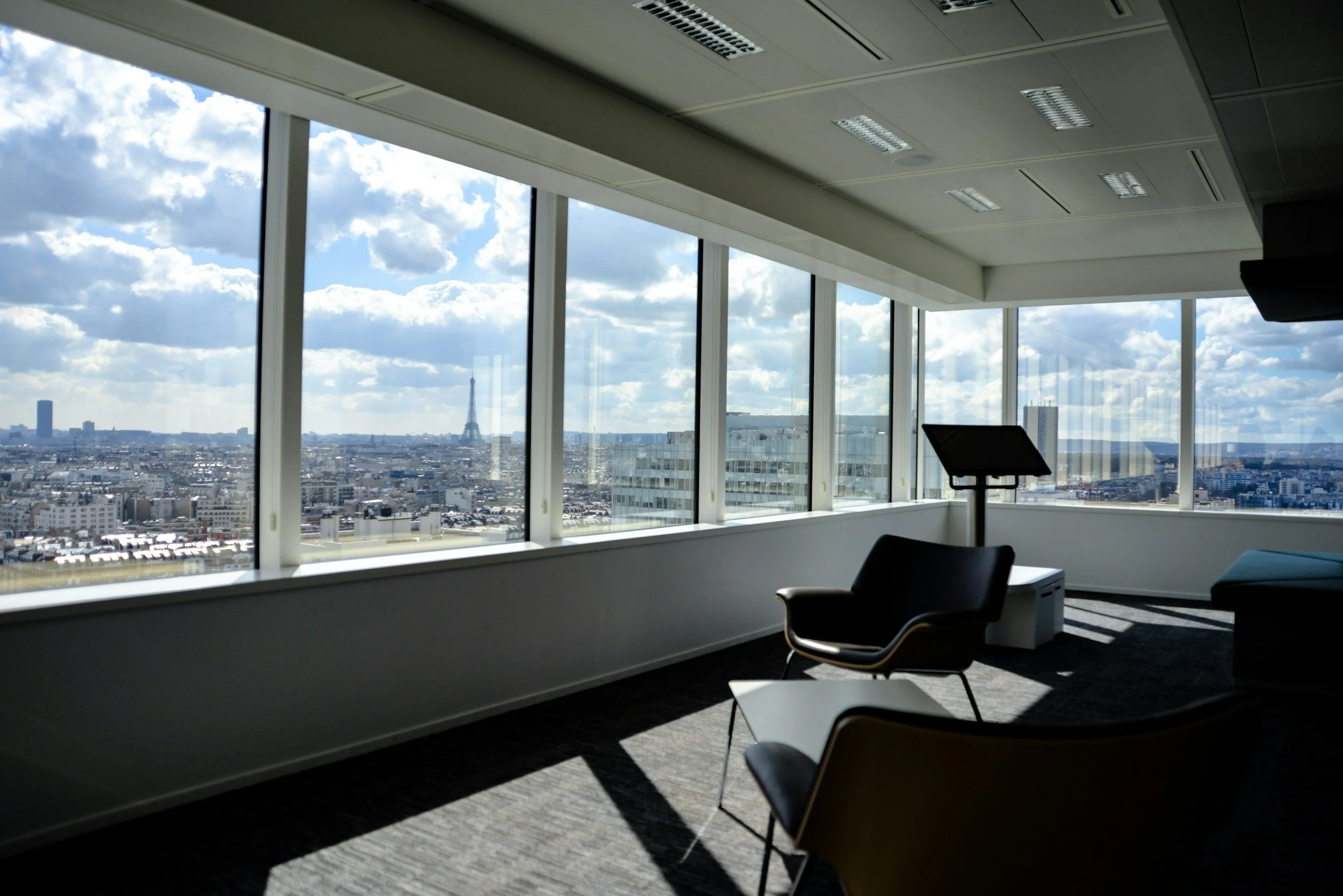 Interior of an office with large windows showing a city skyline with the Eiffel Tower in the distance, seating area with chairs and a tablet stand.