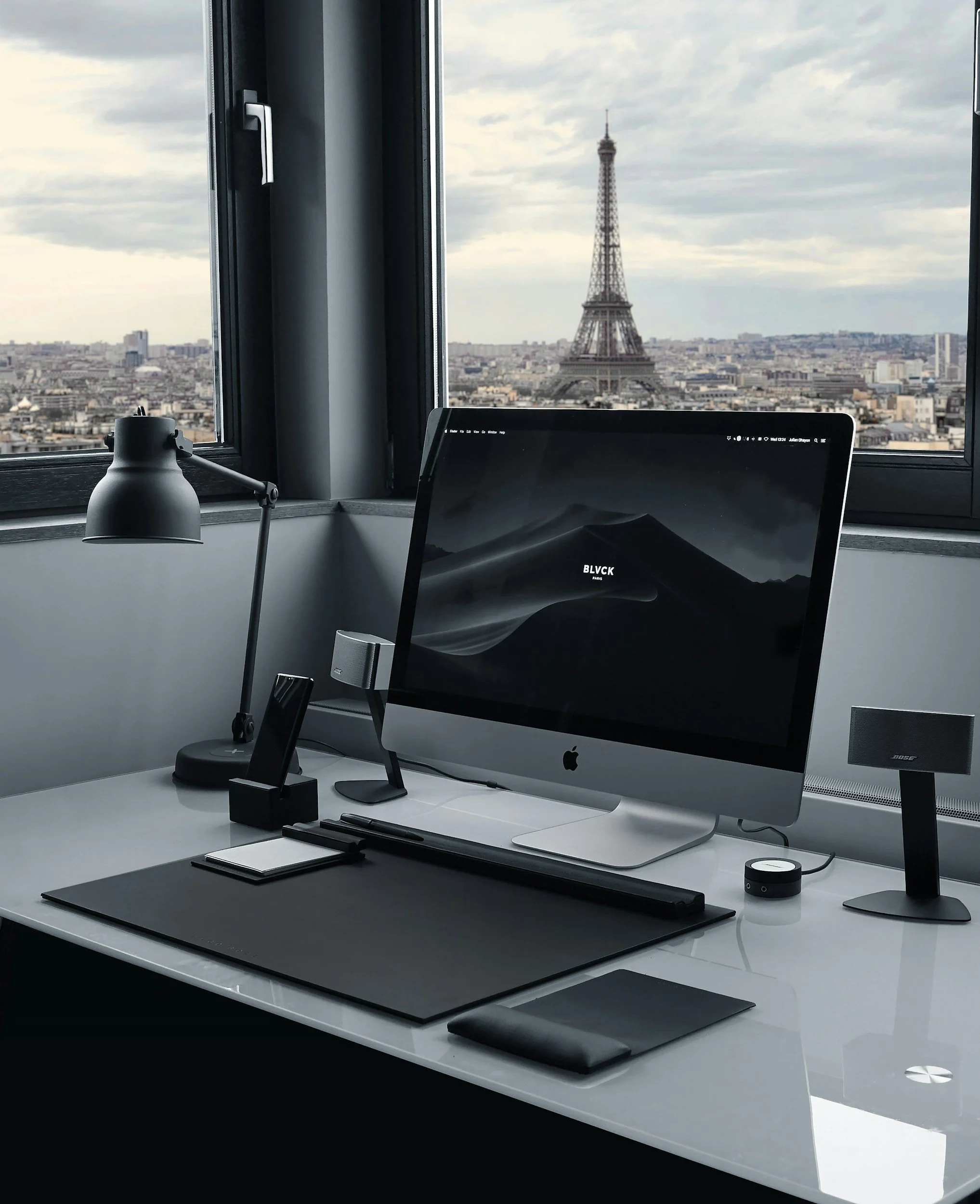 A modern workspace with a white desk, Apple iMac computer, desk lamp, Bose speaker, a small notebook, and a French window with a view of Paris, including the Eiffel Tower in the background.