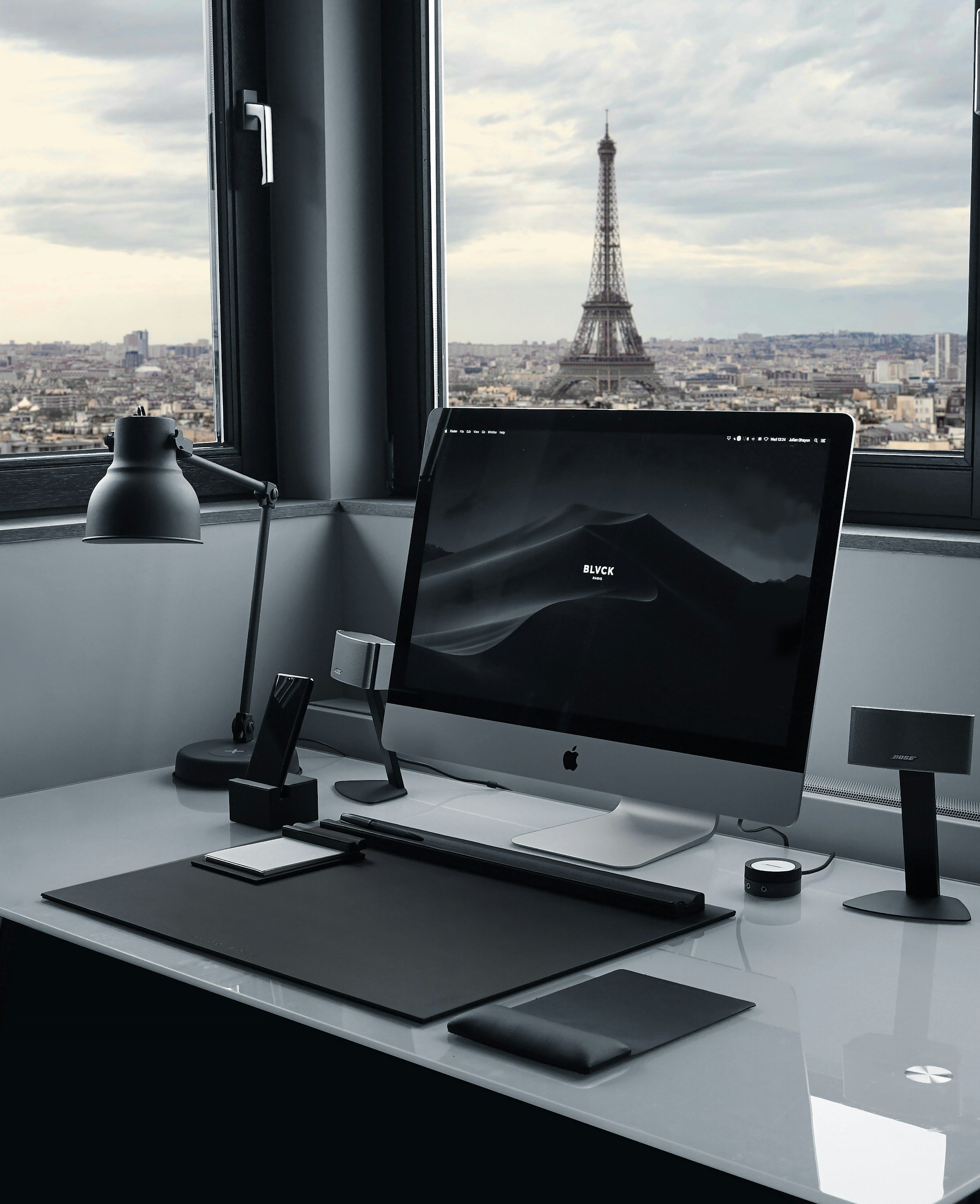 Modern workspace with an iMac computer on a white desk, overlooking the Eiffel Tower in Paris through a large window.