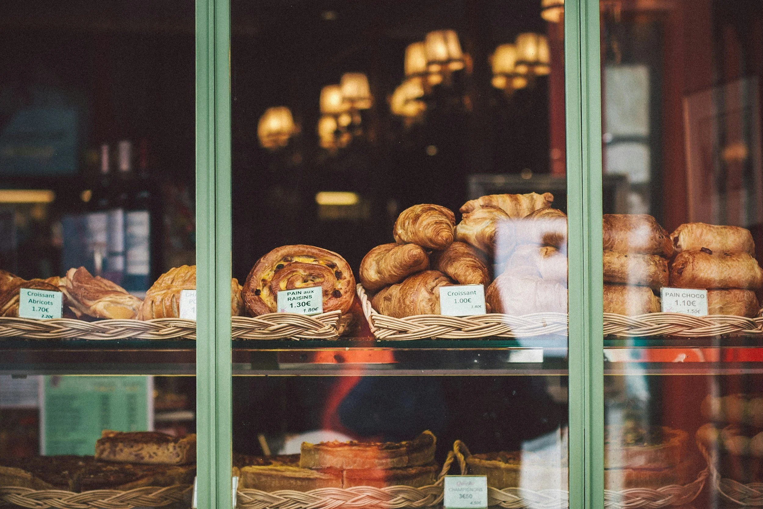 Display window of a bakery showcasing various pastries, including croissants, a raisin pie, and other baked goods, inside the shop.