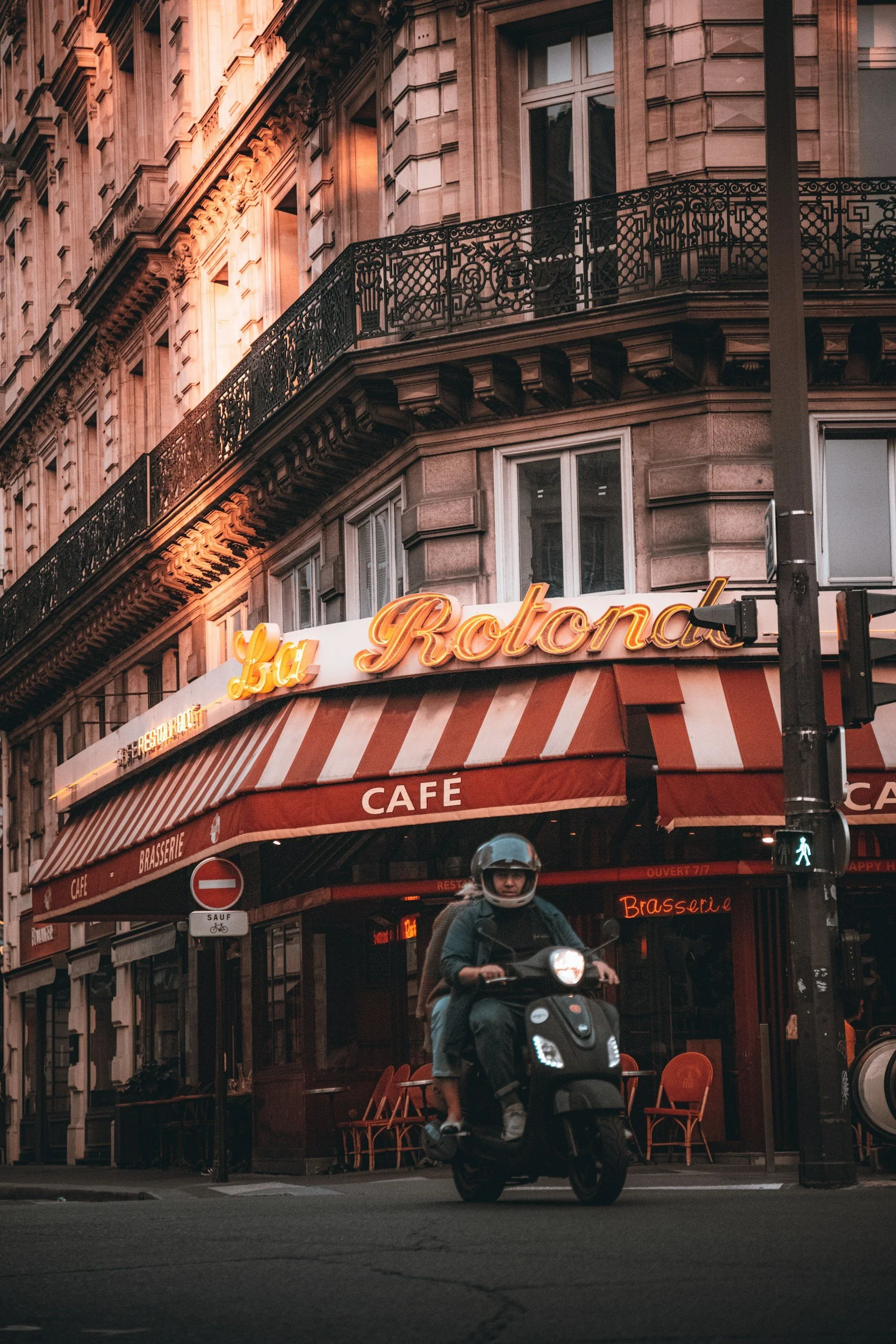 A person riding a scooter in front of a café called La Rotonde in Paris, France, with historic building architecture and a pedestrian crossing signal visible.
