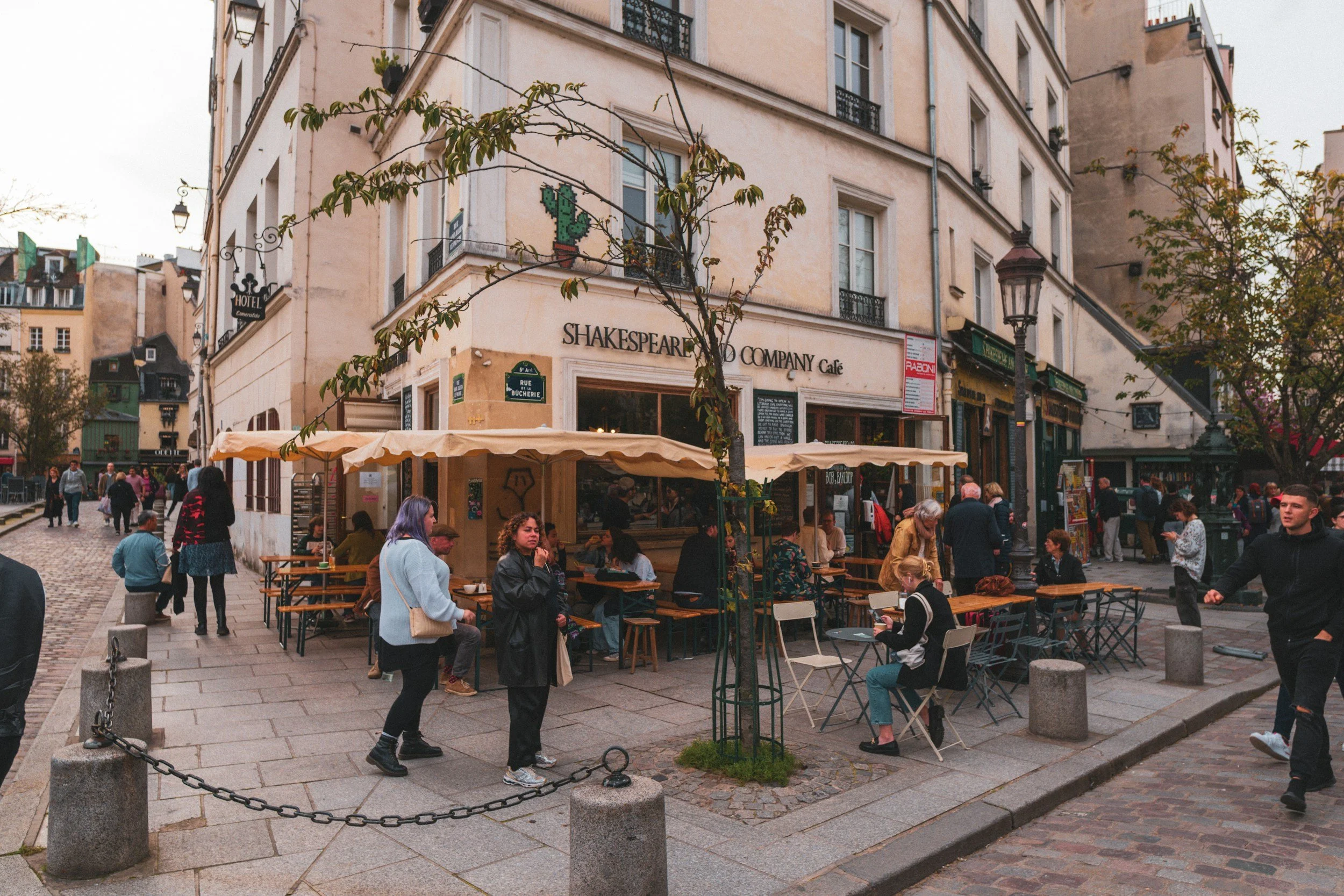 People gathered outside a cafe called Shakespeare and Company Cafe in a Parisian neighborhood with cobblestone streets and surrounding buildings.