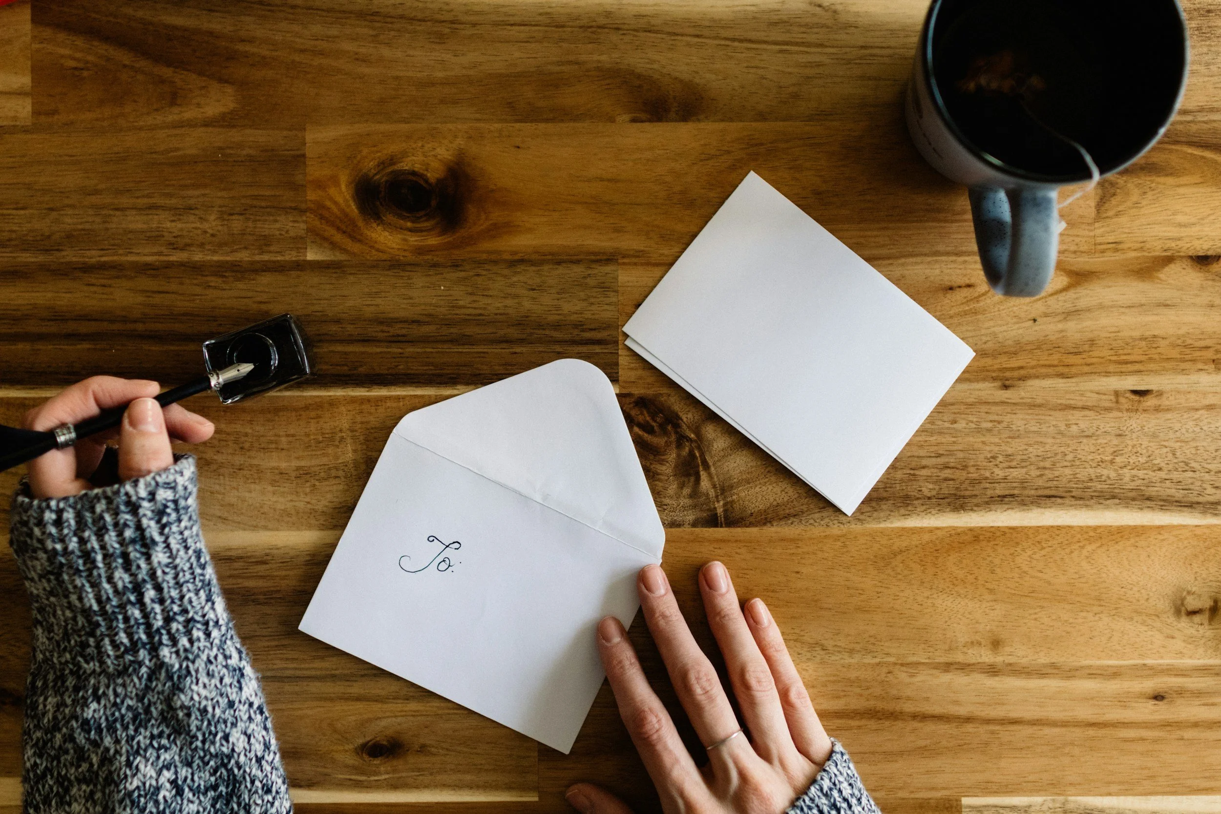 Person in a knit sweater writing 'To:' on a white envelope with a black pen, on a wooden table. There are blank cards, a mug, and a hand with a wedding ring.