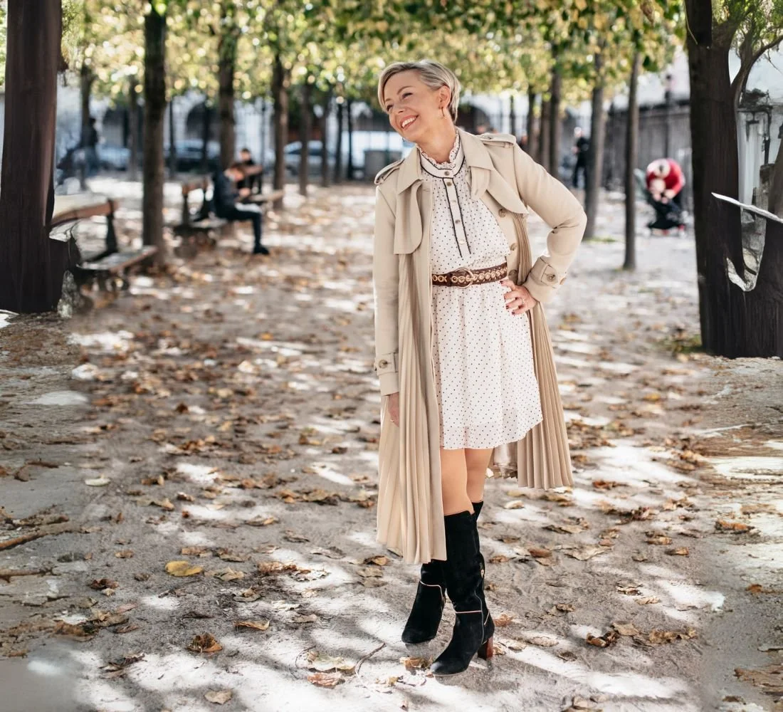 A woman smiling and posing in a park with fallen leaves, wearing a beige trench coat, a white polka-dot dress, and black boots, with trees and benches in the background.