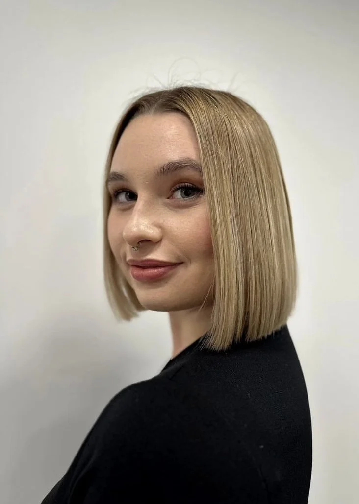 A young woman with shoulder-length blonde hair, light skin, and a septum piercing, wearing a black top, looking over her shoulder with a slight smile against a plain light-colored background.