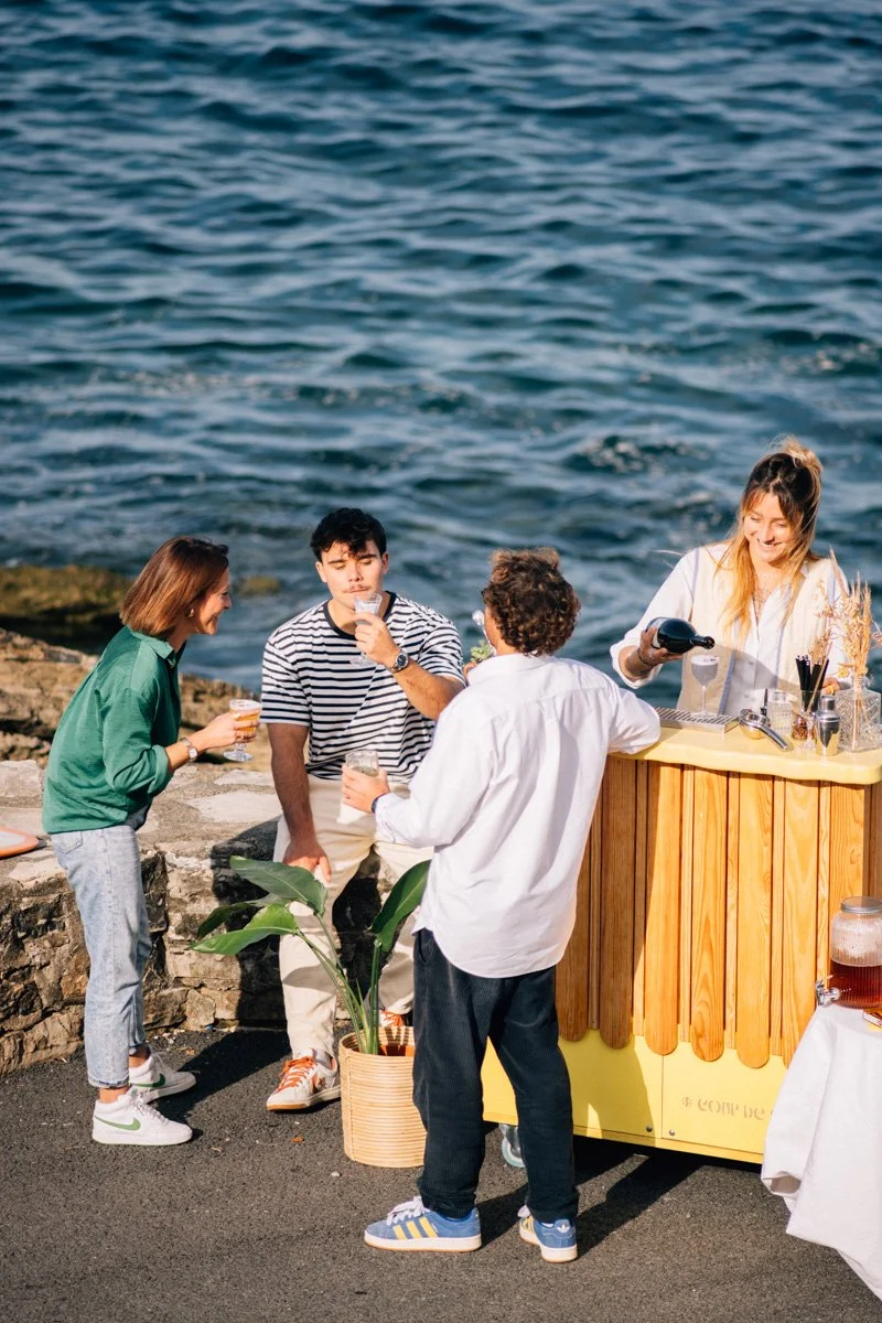 Groupe de quatre personnes profitant d'une dégustation de boissons près de la mer, avec une barmaid derrière un stand en bois, en plein air.