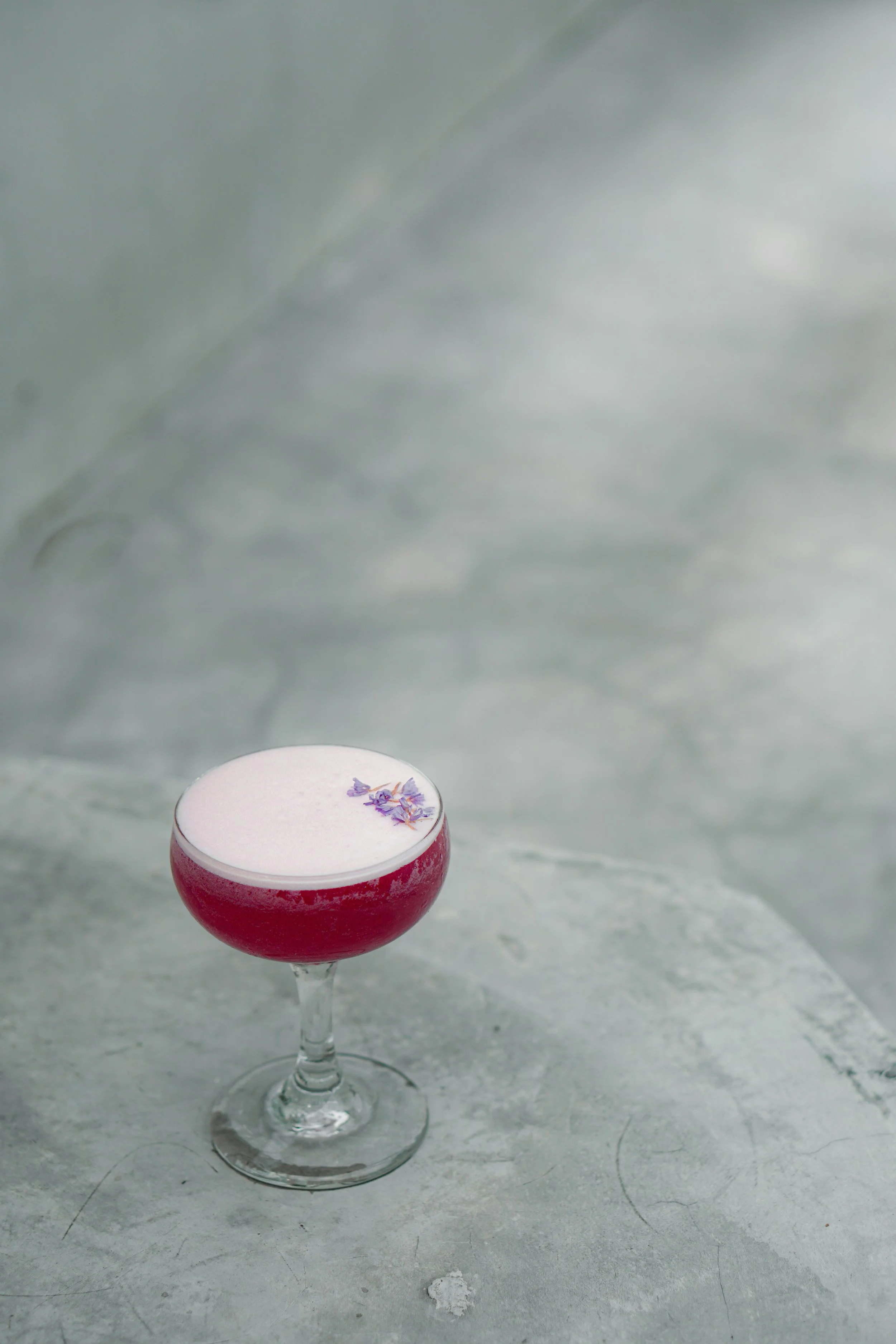 Un cocktail rose avec de la mousse blanche et une garniture florale sur le dessus, posé sur une surface grise.