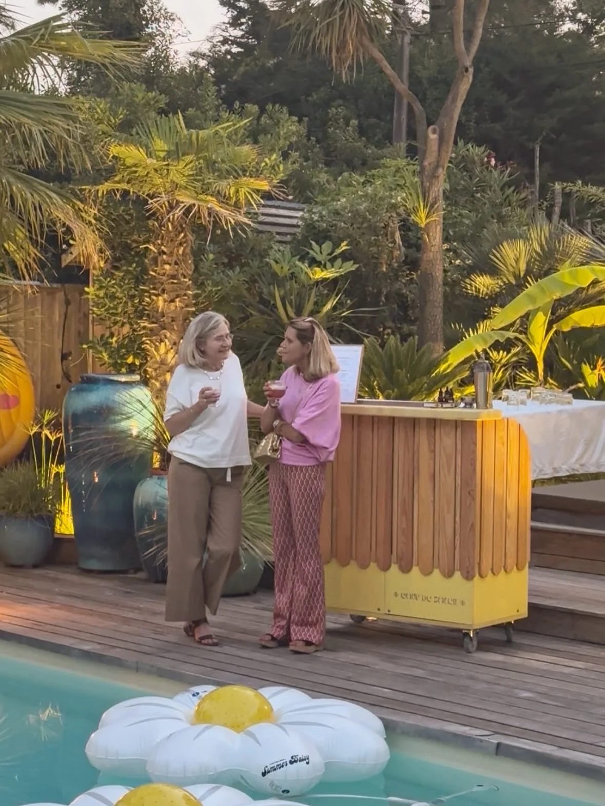 Deux femmes discutent près d'un bar en bois dans un espace extérieur avec beaucoup de végétation tropicale, en train de tenir des verres avec une boisson. Il y a des ballons flottants dans la piscine à l'avant-plan.