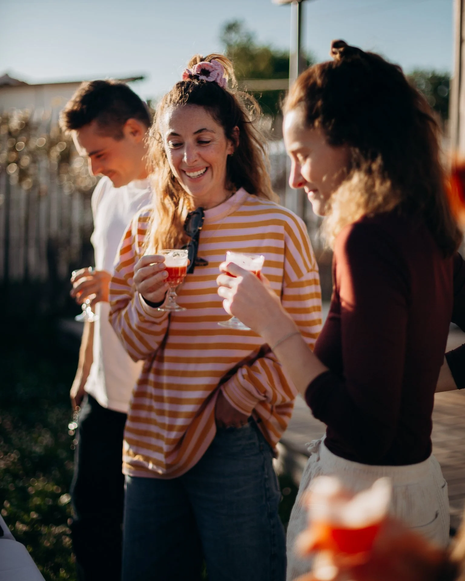 Groupe de personnes souriantes qui tiennent des verres avec des boissons, profitant d'une réunion en plein air lors d'une journée ensoleillée.