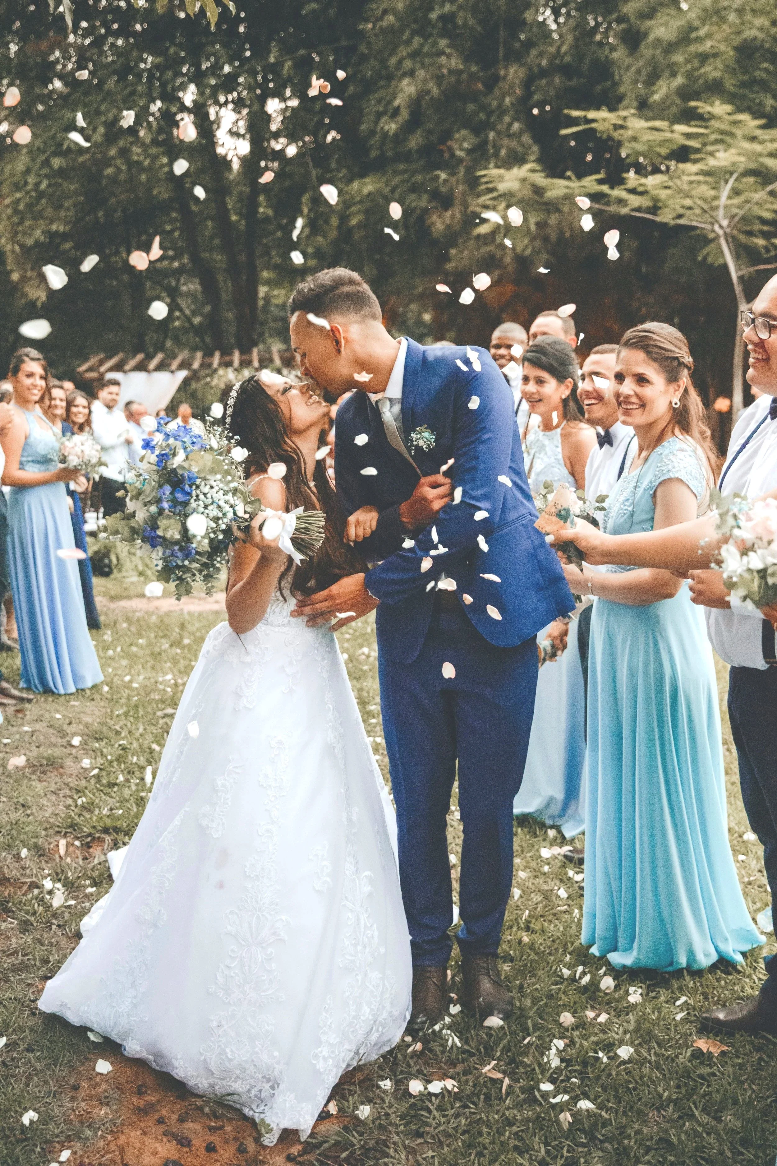 Un couple de mariés partage un baiser lors d'une cérémonie de mariage en plein air, entourés de témoins et d'amis, avec des pétales de fleurs volant dans l'air.