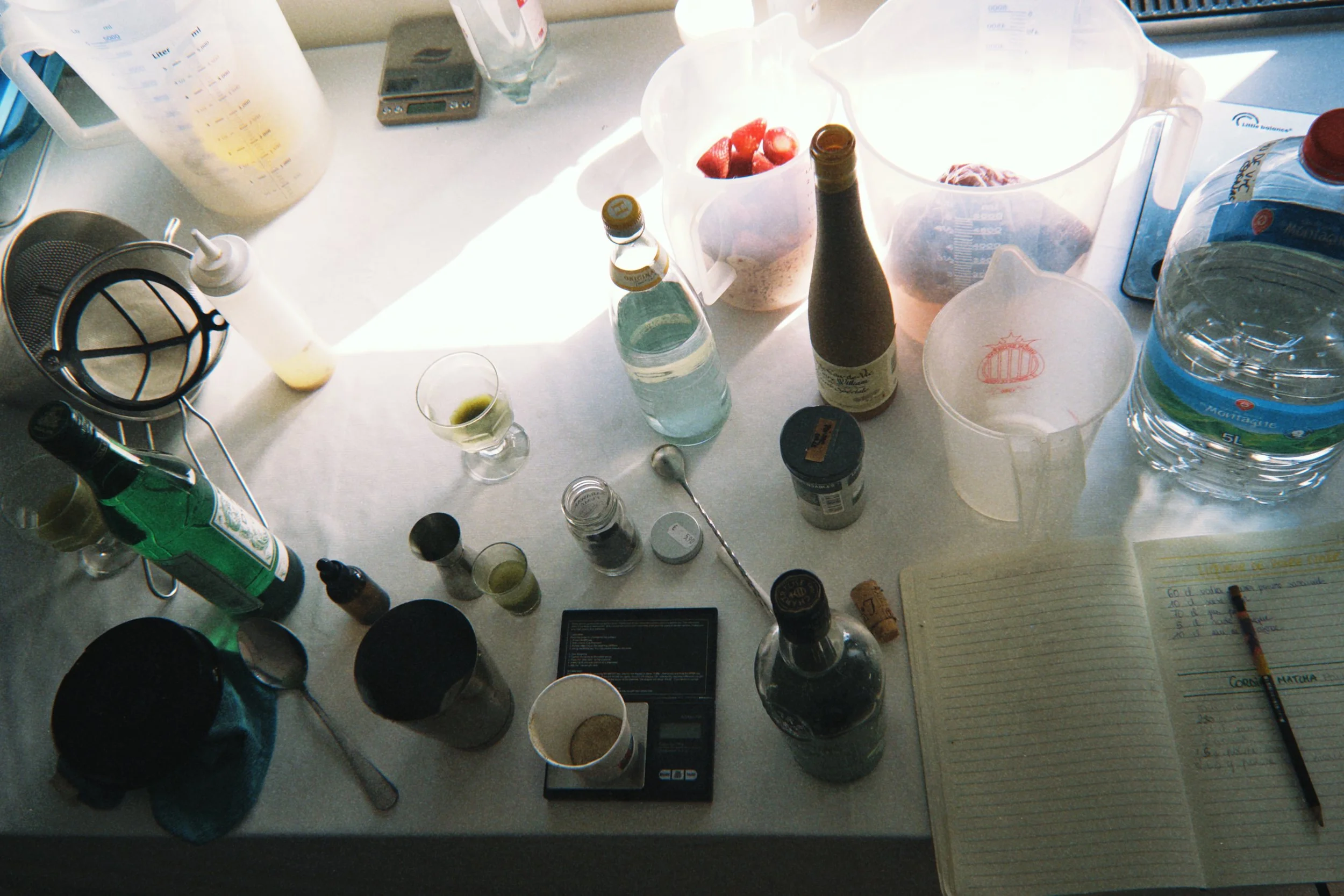 Table de cuisine avec divers ingrédients et ustensiles, comprenant des bouteilles, des pommes de terre, une balance, un carnet de notes, un shaker à cocktail, et d'autres contenants transparentes, sous une lumière naturelle. fait maison