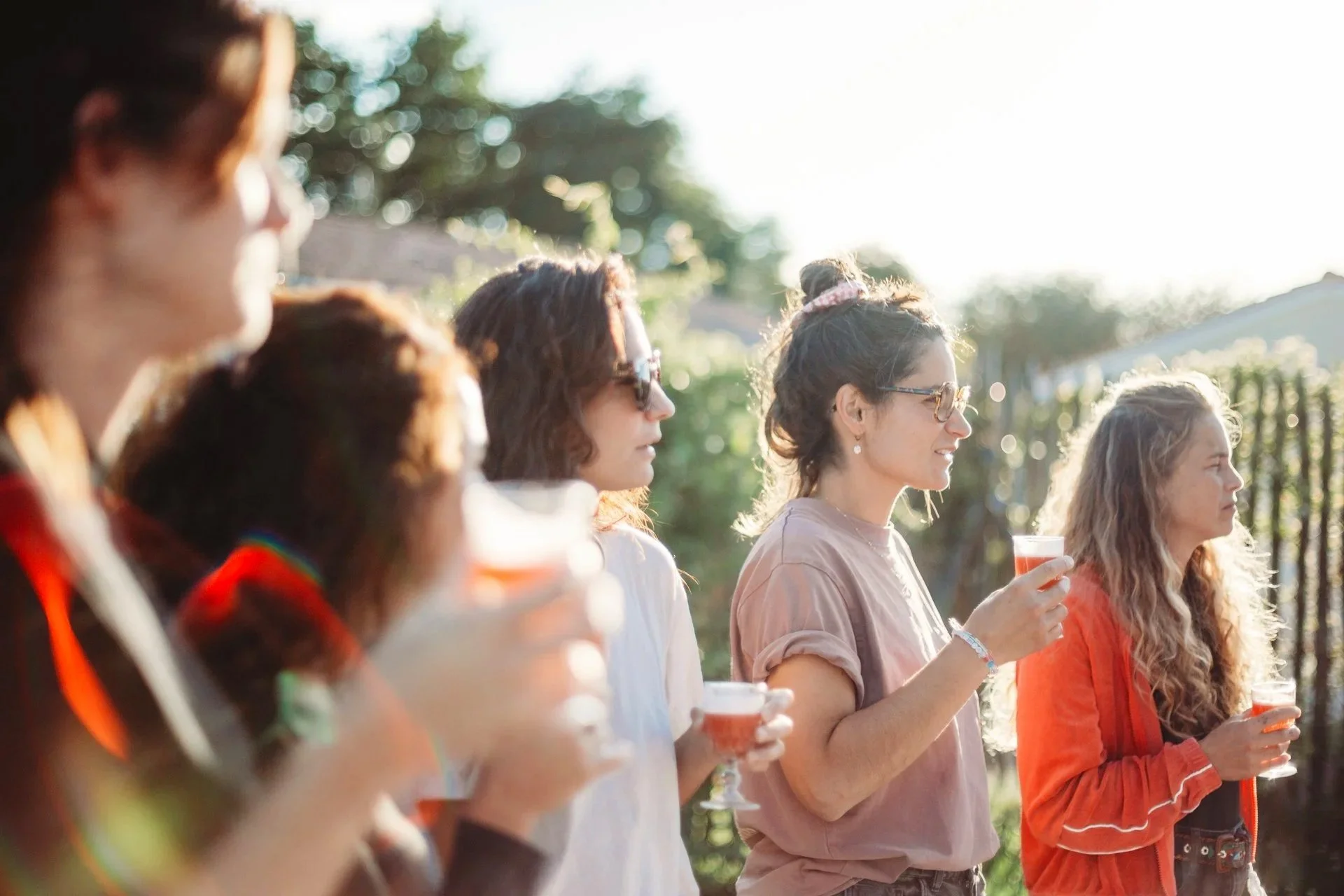 Groupe de personnes lors d'une fête ou rassemblement en extérieur, tenant des verres de boisson, avec éclairage naturel et arrière-plan d'arbres.