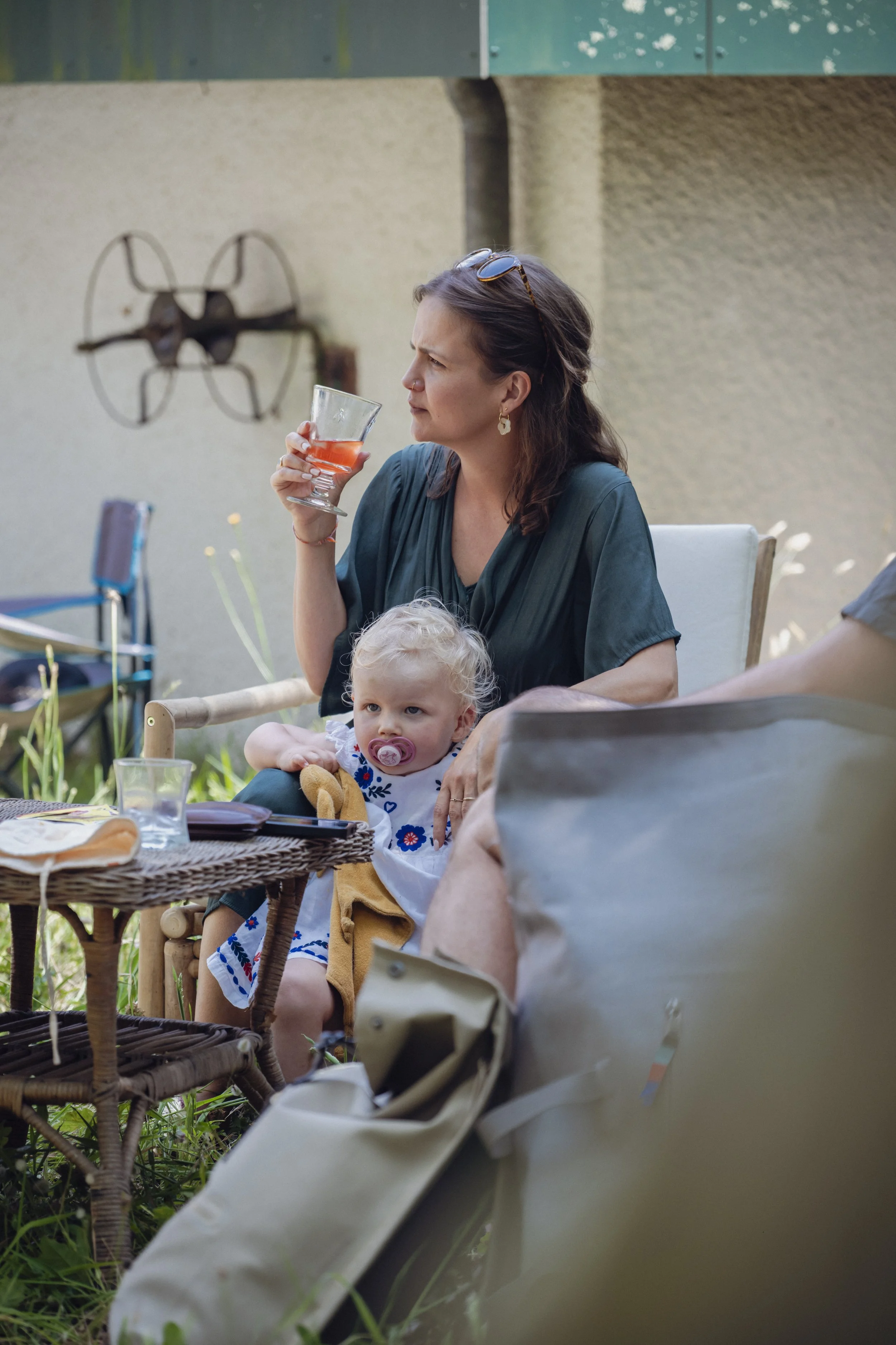 Une femme assise à une table extérieure, tenant un verre avec une boisson rose, avec un enfant assis sur ses genoux. La femme porte des lunettes sur la tête et des boucles d'oreilles, tandis que l'enfant a une sucette dans la bouche. Il y a des objets sur la table, comme des verres et des assiettes, et un fond de mur beige avec une pièce de décoration métallique en forme d'avion.