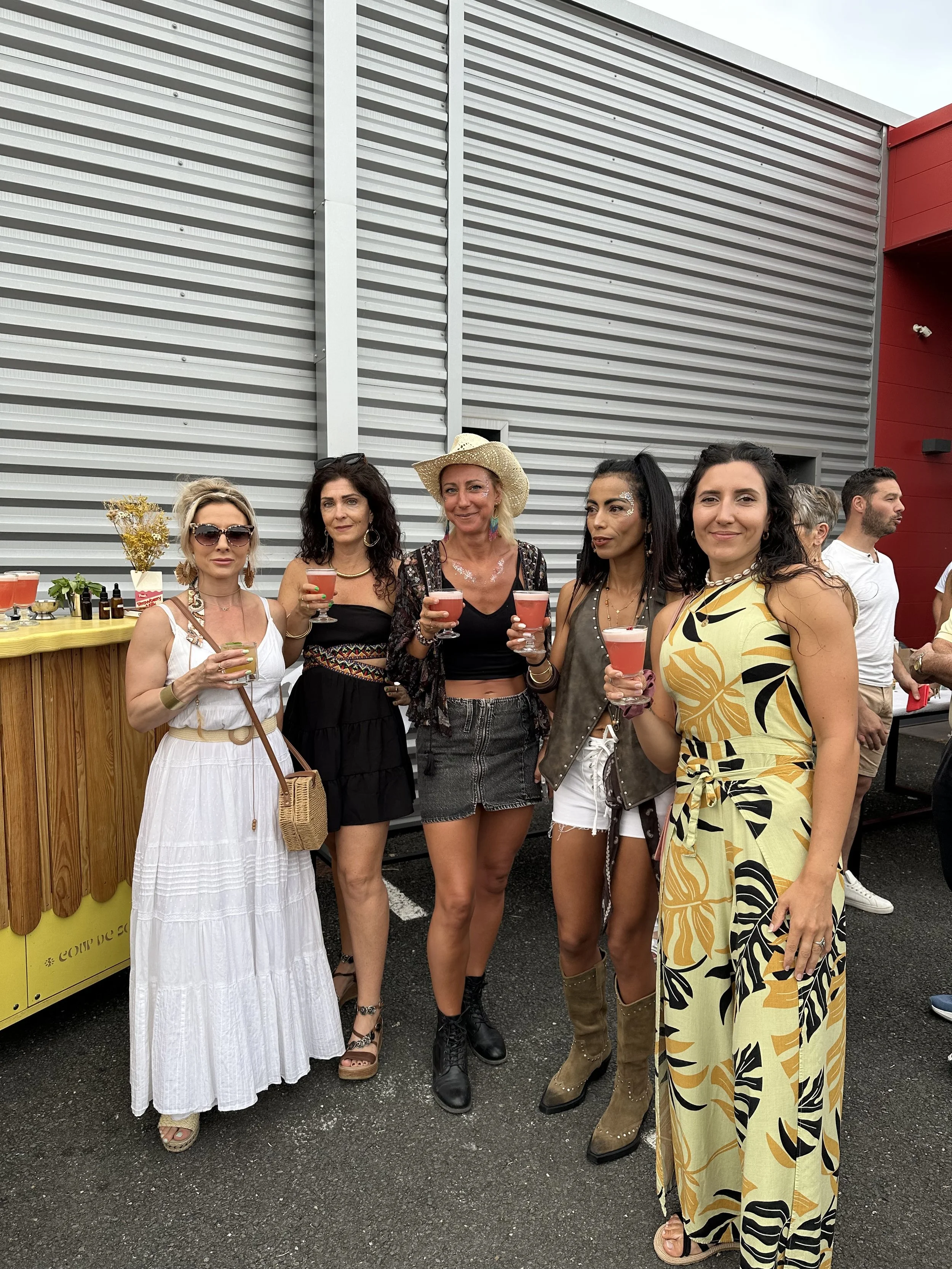 Un groupe de femmes posant avec des verres de cocktail lors d'une fête en extérieur, devant un mur en métal et un comptoir en bois.