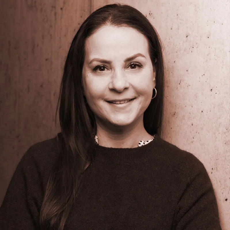A woman with dark brown hair smiling, wearing a black sweater and a polka-dot shirt underneath, standing against a textured wall.