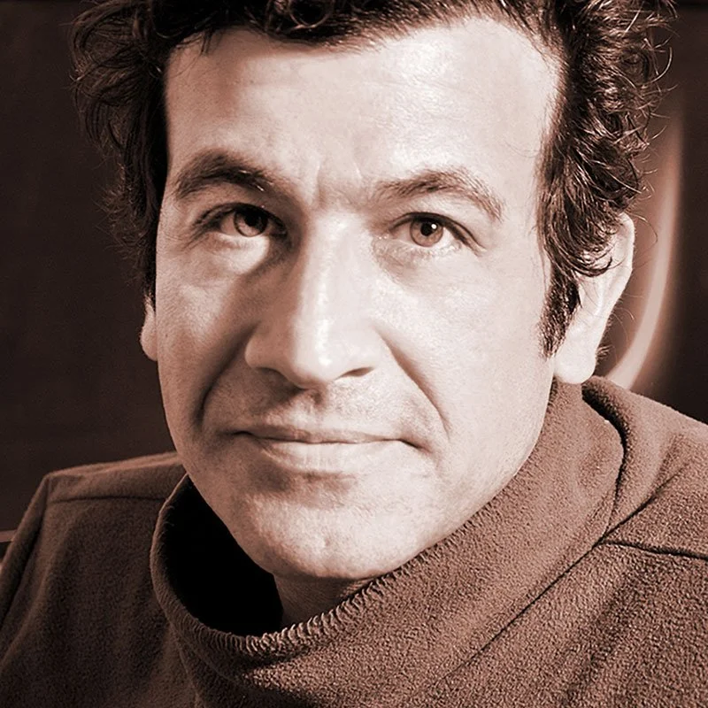 Close-up of a man with curly dark hair and light skin, wearing a brown turtleneck sweater.