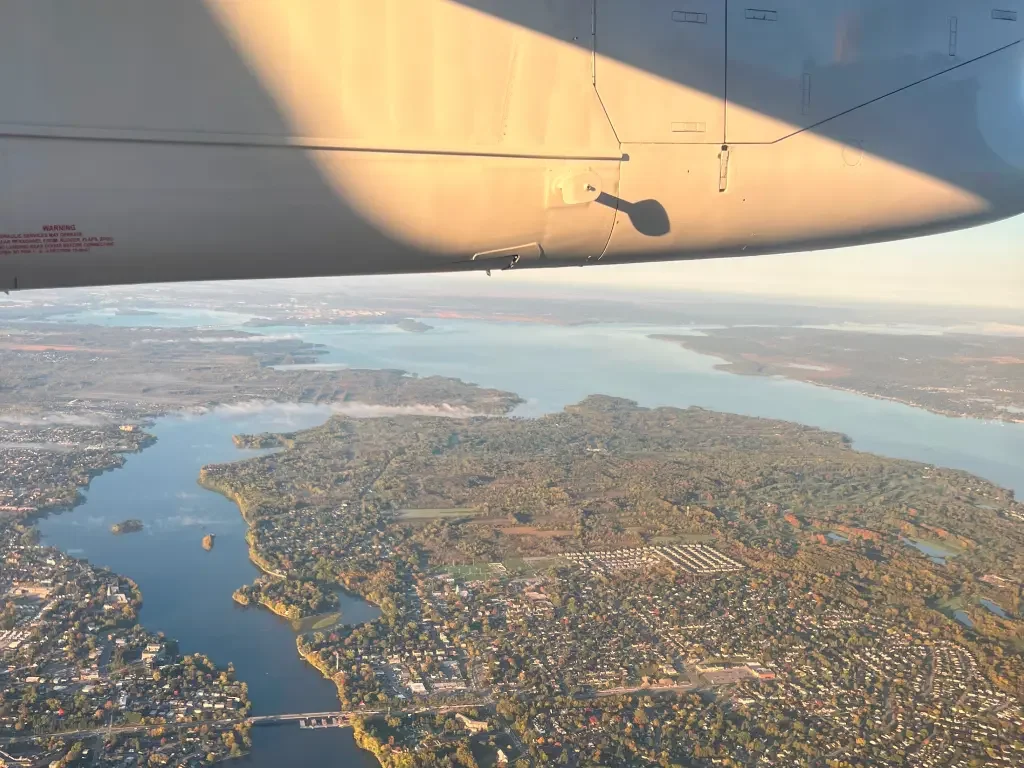 Aerial view from a plane showing forests, lakes, and visible communities on the ground, with part of the aircraft wing in frame.