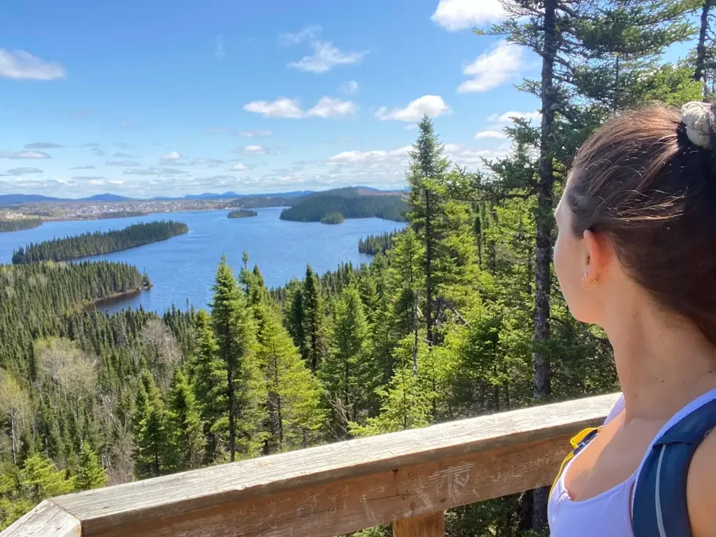 Hiking in Oujé-Bougoumou, a woman travel nurse admires panoramic views of a vast lake and boreal forest under a sunny blue sky from a lookout.