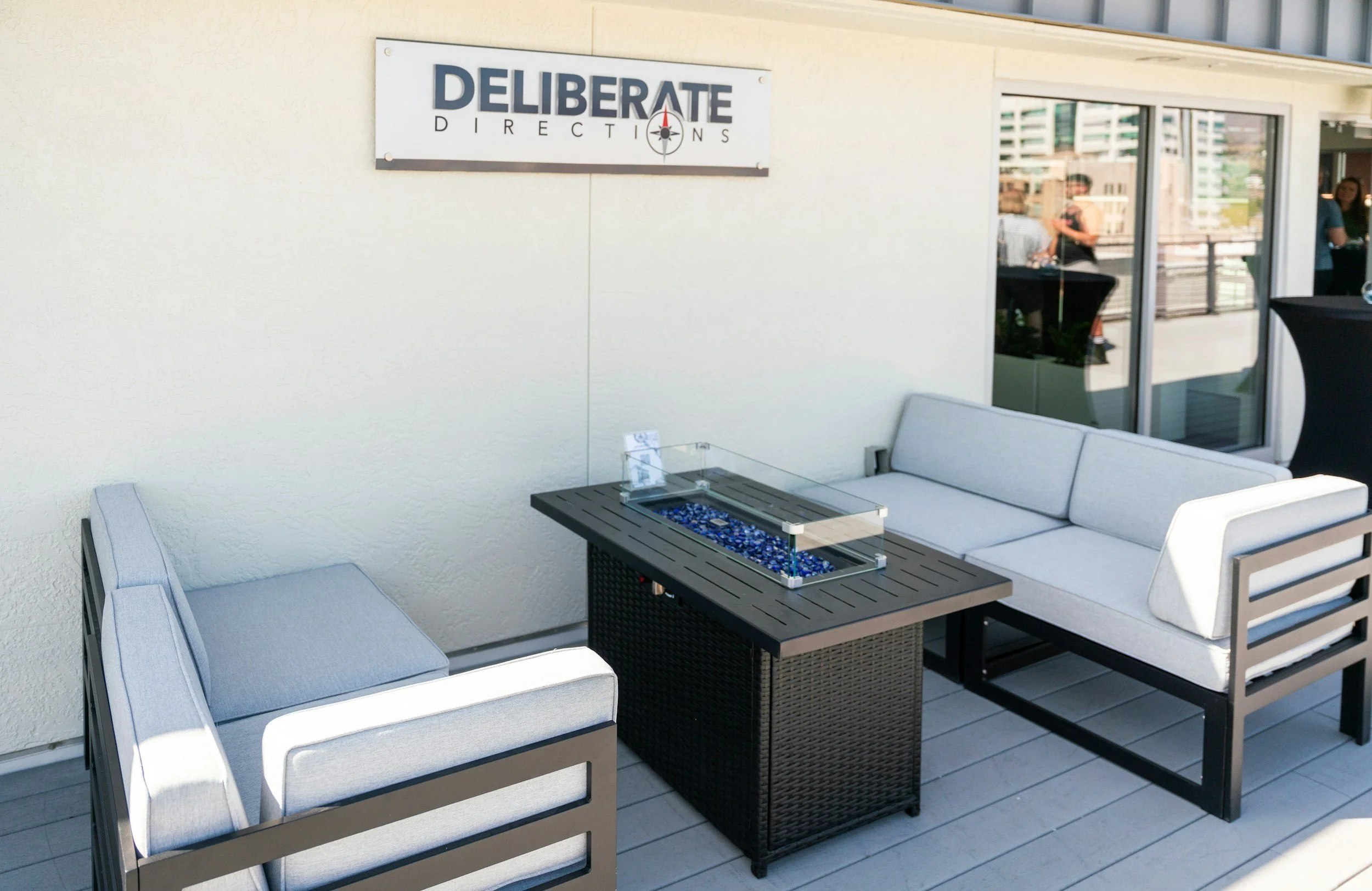 Outdoor patio seating area with a sign that reads "Deliberate Directions", modern cushioned chairs and a table with a glass box filled with blue glass beads near a window.