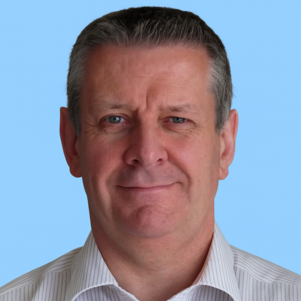 Close-up of a middle-aged man with short, gray hair and blue eyes, smiling slightly, wearing a white collared shirt, against a blue background.