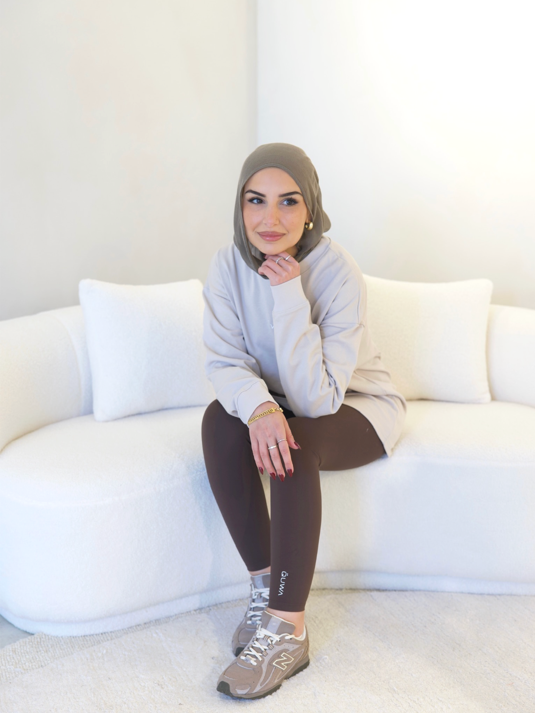 A woman wearing a beige hoodie, brown leggings, and gray sneakers sitting on a white couch in a minimalist room with cream-colored walls.