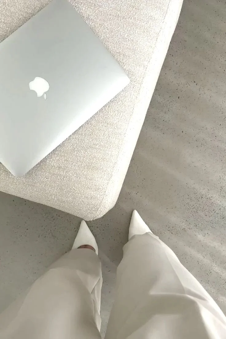 View of a person standing on a light gray concrete floor, wearing white pointed-toe shoes and white pants, taken from a high angle. There is a beige upholstered bench with a closed silver MacBook laptop resting on it.