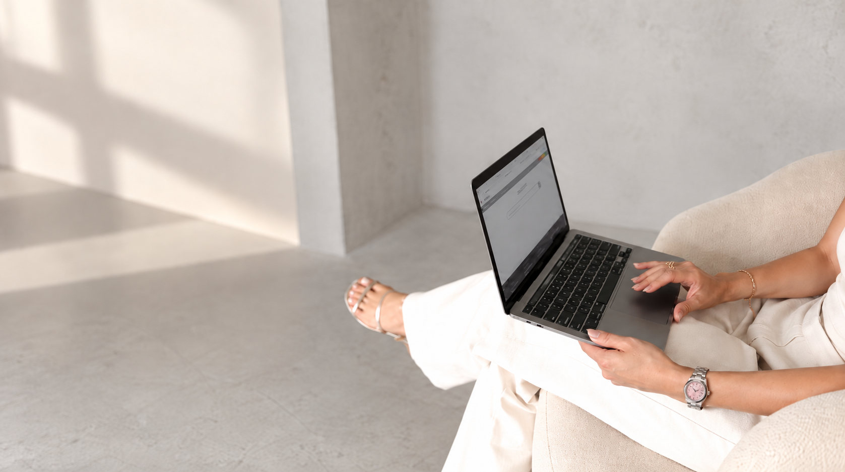 A person sitting on a light-colored sofa using a laptop, with legs crossed and wearing white pants, a watch, and jewelry, in a minimalistic room with a concrete or plaster wall.