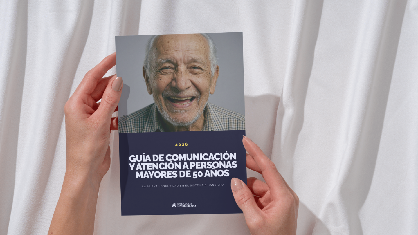 Persona mayor sonriendo sosteniendo una guía de comunicación para personas de más de 50 años frente a una cortina blanca.