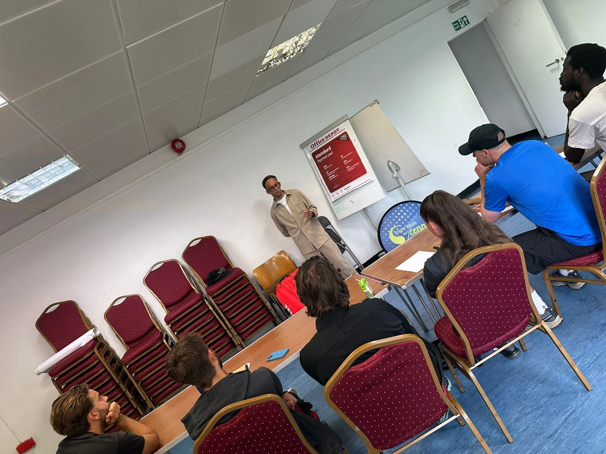 A classroom setting with students seated at tables, some with laptops and notebooks. A woman is standing at the front, possibly instructing. There is a sign on the wall titled 'Office Depot' indicating a workshop or class related to office supplies.