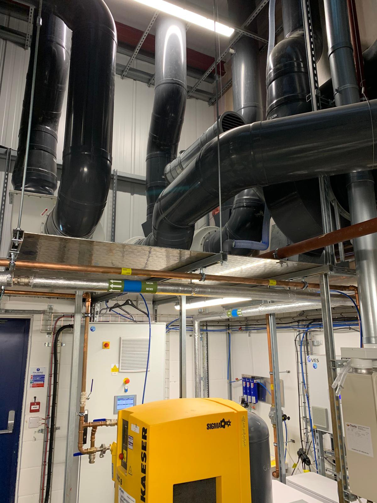 Industrial room containing black ventilation ducts, a yellow air compressor labeled 'KAESER,' pipes, electrical wiring, and a white control panel.