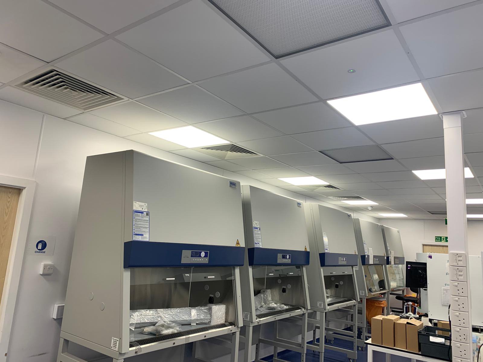 A laboratory with multiple fume hoods along one wall, various boxes, and a computer workstation, under fluorescent lighting.