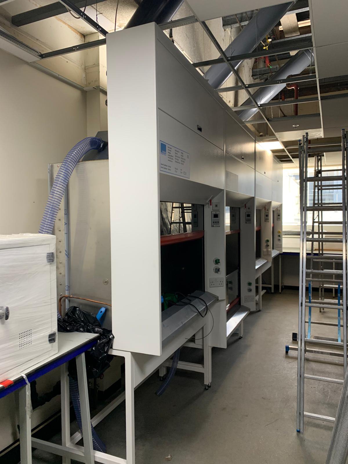 Laboratory fume hoods and equipment in an industrial setting with exposed ceiling pipes and a metal ladder.