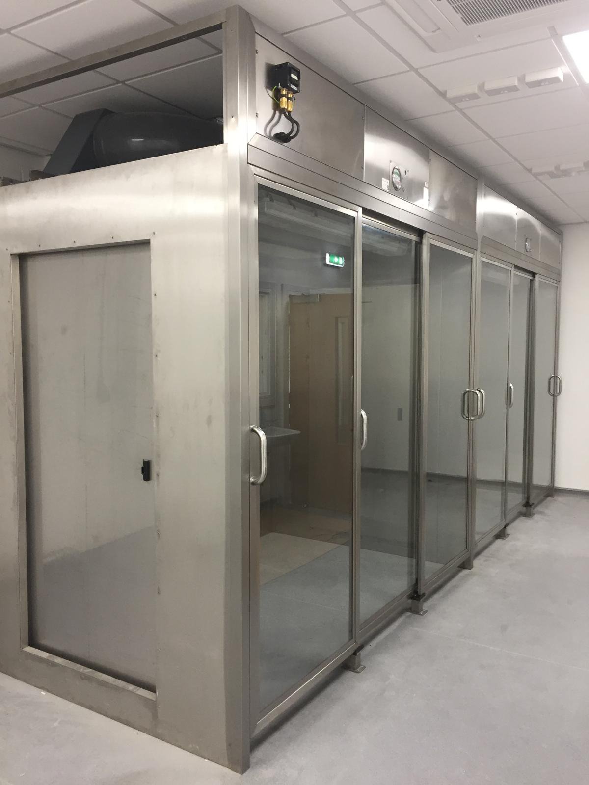 A large metallic industrial walk-in refrigeration unit with glass doors and handles, inside a facility with a tiled ceiling and concrete floor.