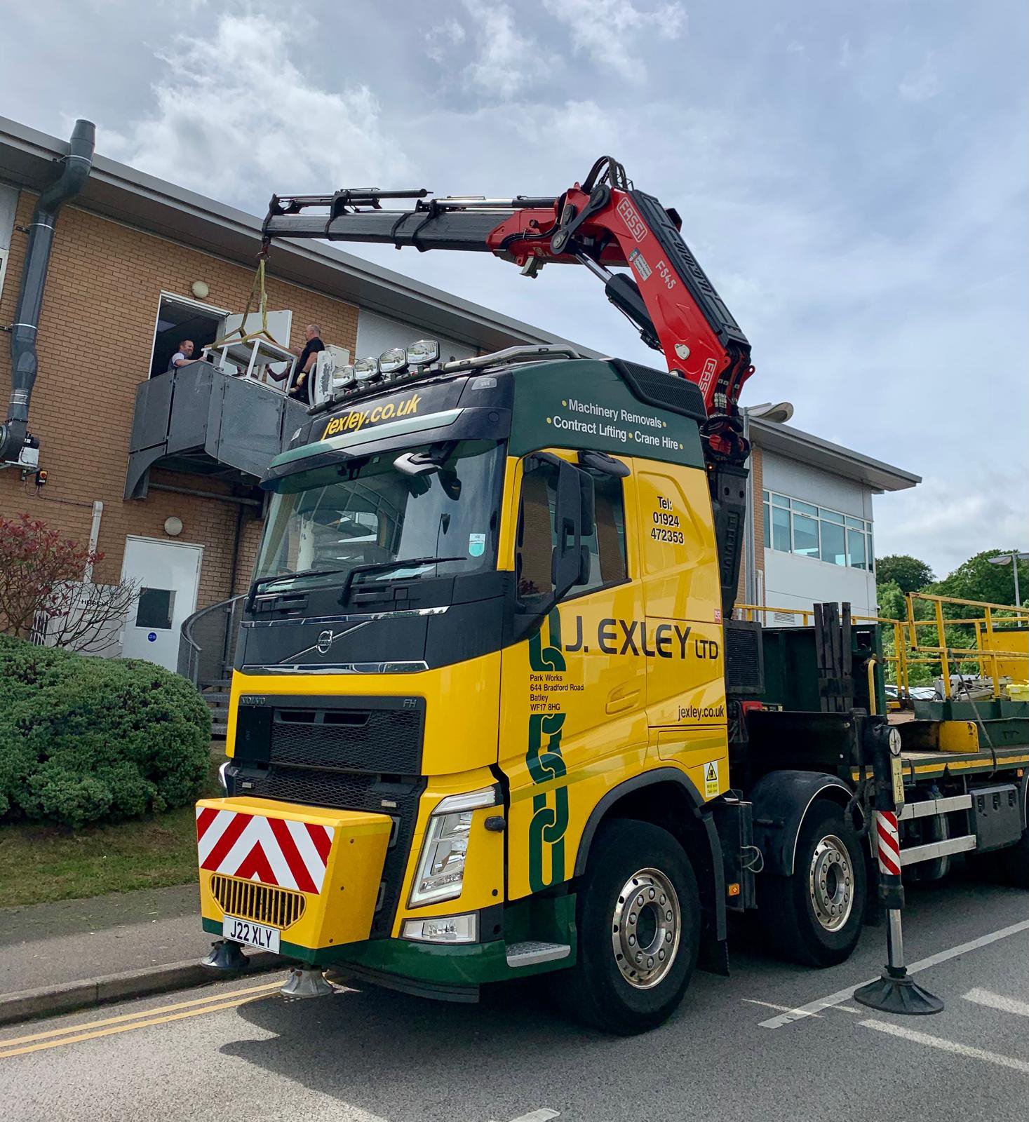 A large yellow and green truck with the logo 'J. Exley Ltd' is positioned beside a building. The truck has an extended crane arm reaching up to a balcony where two people are loading or unloading a white machine or piece of equipment. The truck speci