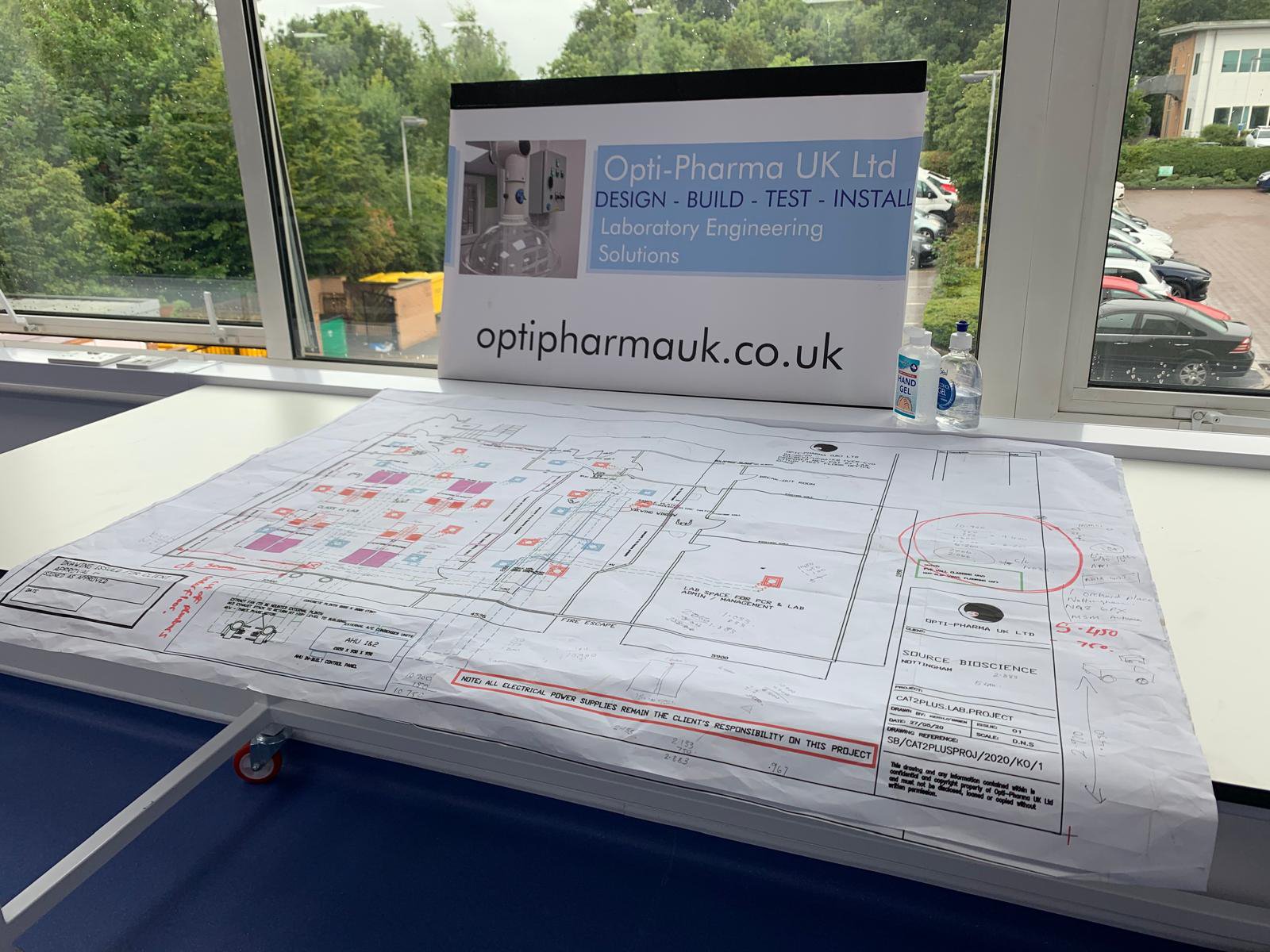Blueprint on a table with "Opti-Pharma UK Ltd" sign, showing laboratory engineering solutions, and a website address. Hand sanitizer bottles nearby.