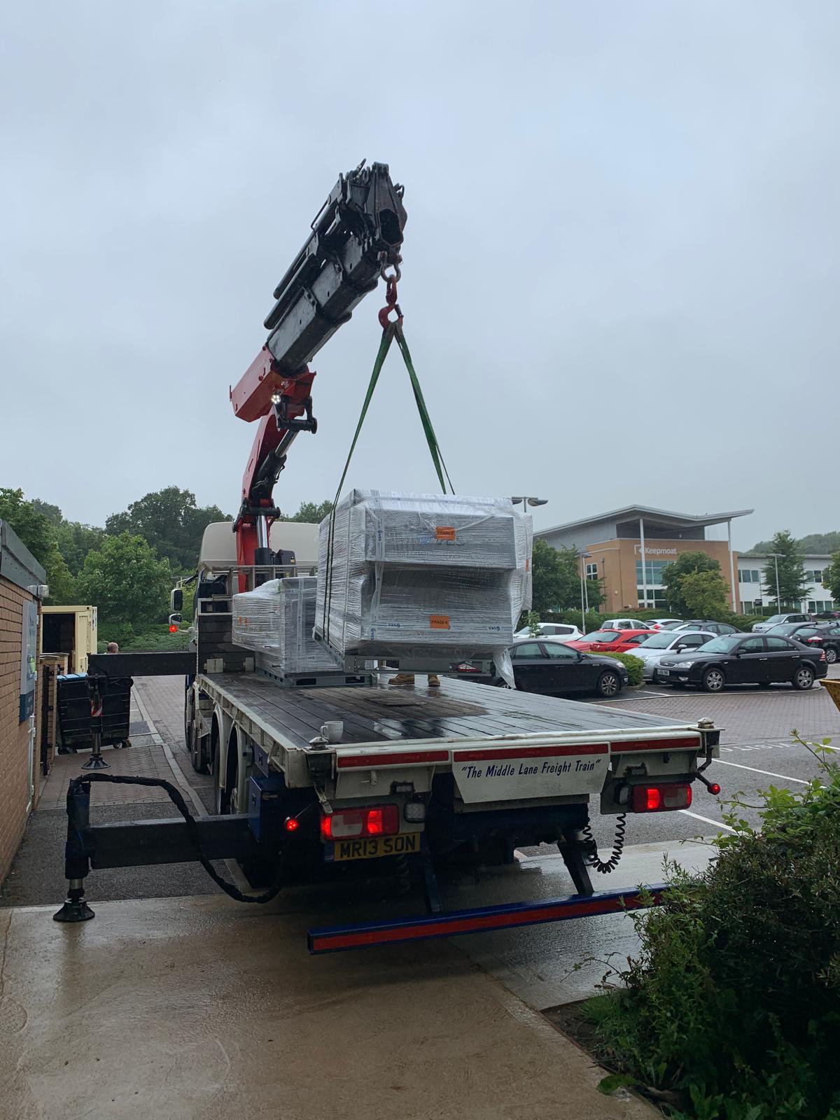 Flatbed truck with crane unloading large wrapped packages in a parking area.