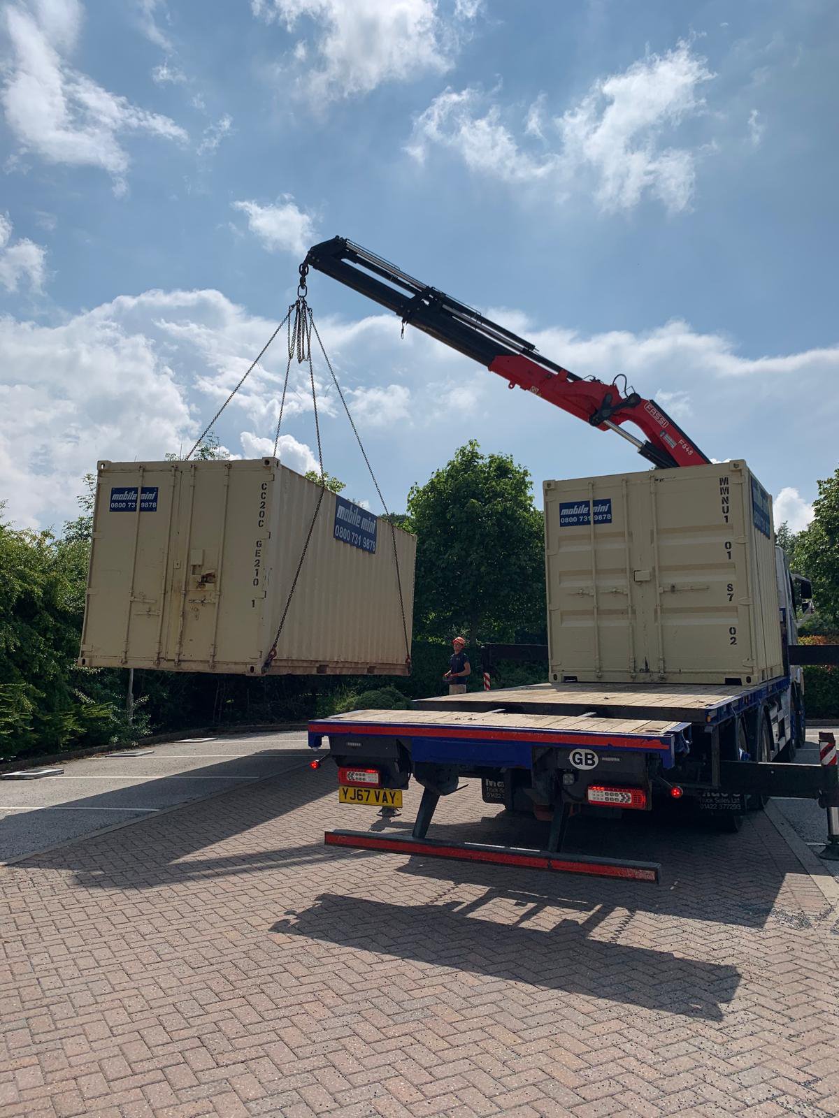 Crane lifting a large beige storage container from a flatbed truck, with a blue sky in the background.