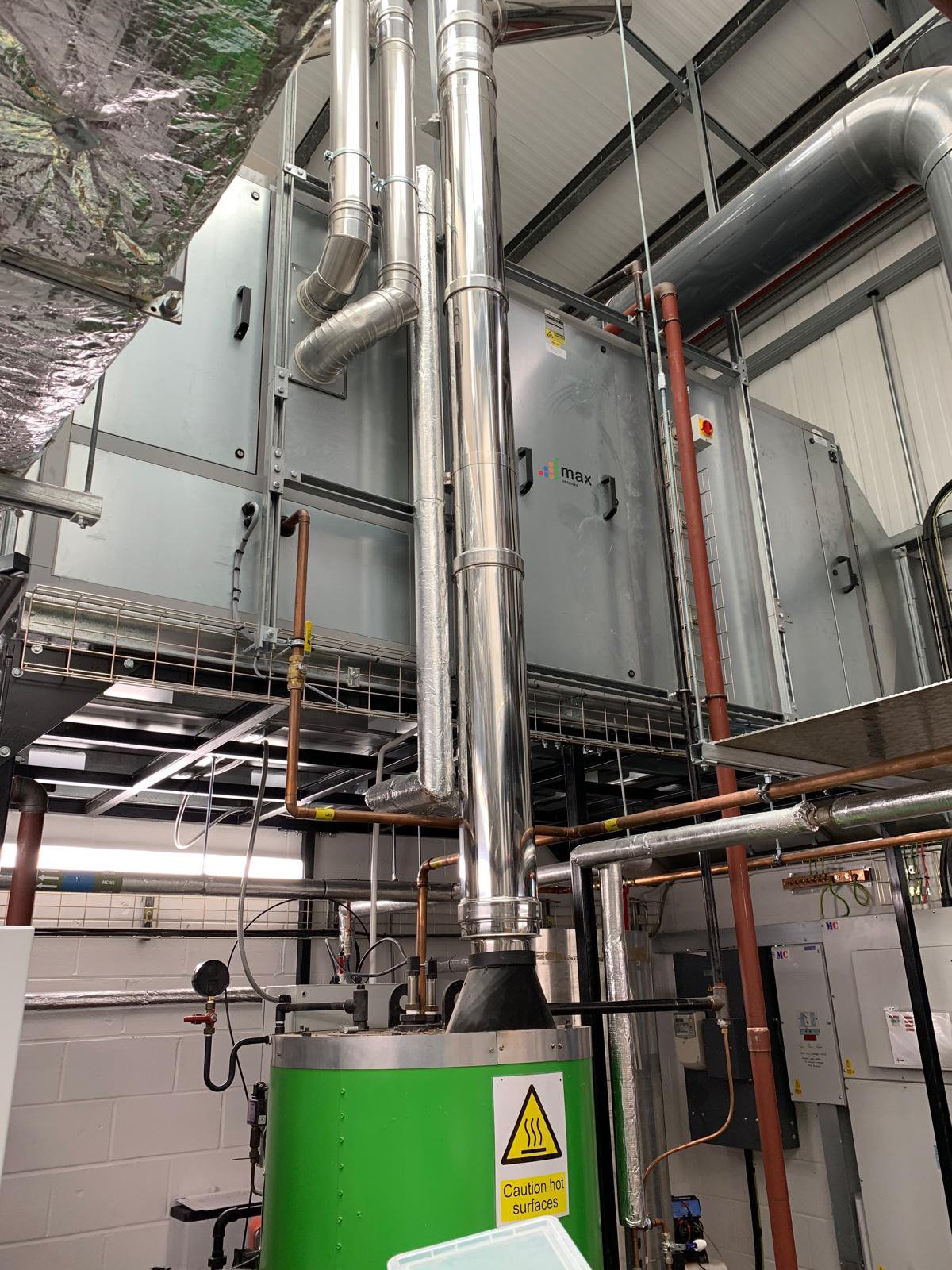 Industrial HVAC system with pipes and a green boiler. Warning sign on the boiler reads "Caution hot surfaces." Pipes and ductwork are visible in a mechanical room.