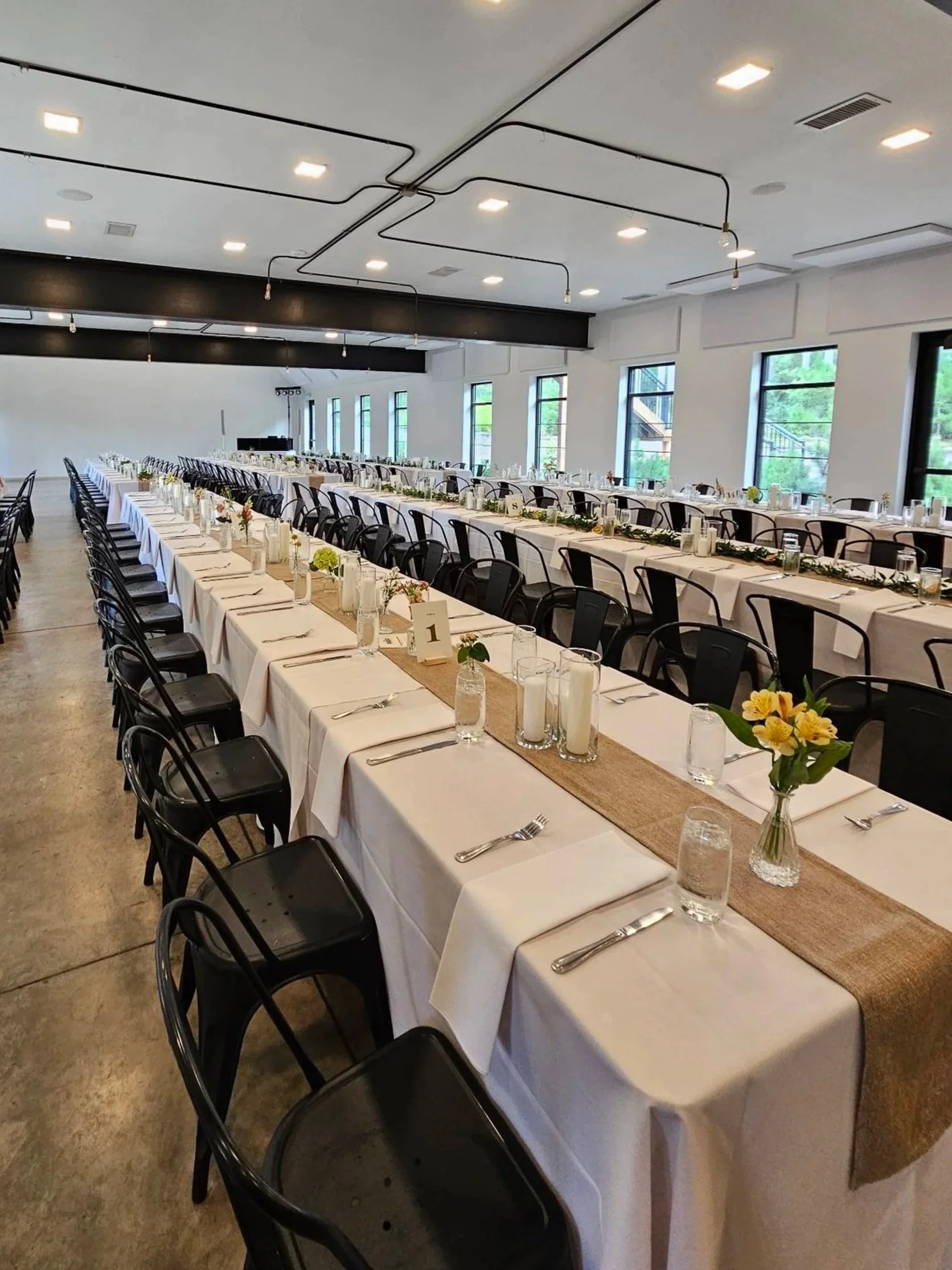 Long banquet tables with white tablecloths, decorated with flowers and candles, set for a formal event in a bright, spacious room with large windows.