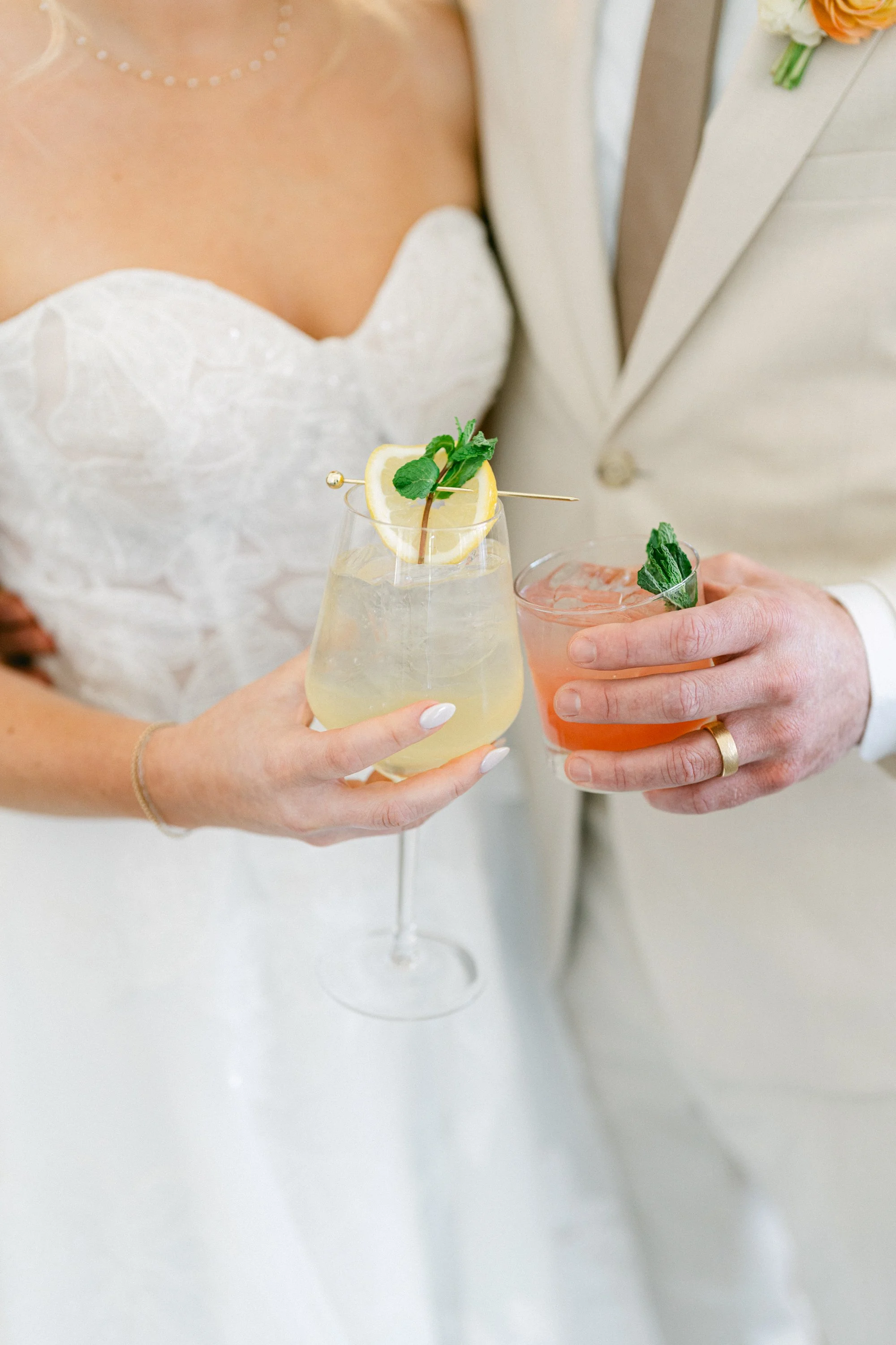 A bride and groom holding cocktails with lemon, mint, and pink garnish, dressed in wedding attire.